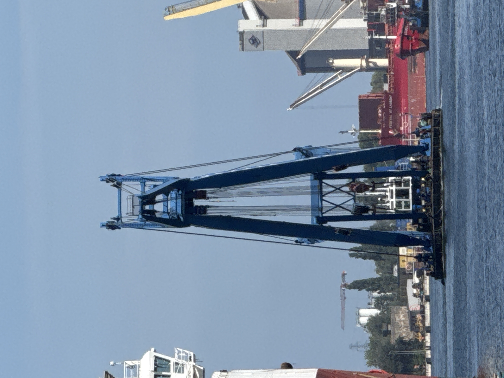 The image depicts a maritime industrial scene, likely within the port of Gdańsk, Poland, under a clear blue sky. Dominating the foreground is a large, blue gantry crane mounted on a floating platform. The crane is intricate, with numerous cables and pulleys visible, suggesting heavy-duty lifting capabilities. To the left, a portion of a white ship with a red hull is visible. In the background, a red cargo ship named "FEDERIK ST LAURENT" is docked. Other industrial structures, including silos and cranes, are present along the waterfront, along with scattered trees and other vessels. A few figures can be seen on the floating platform of the gantry crane, seemingly engaged in operations. The water is calm, reflecting the blue sky.