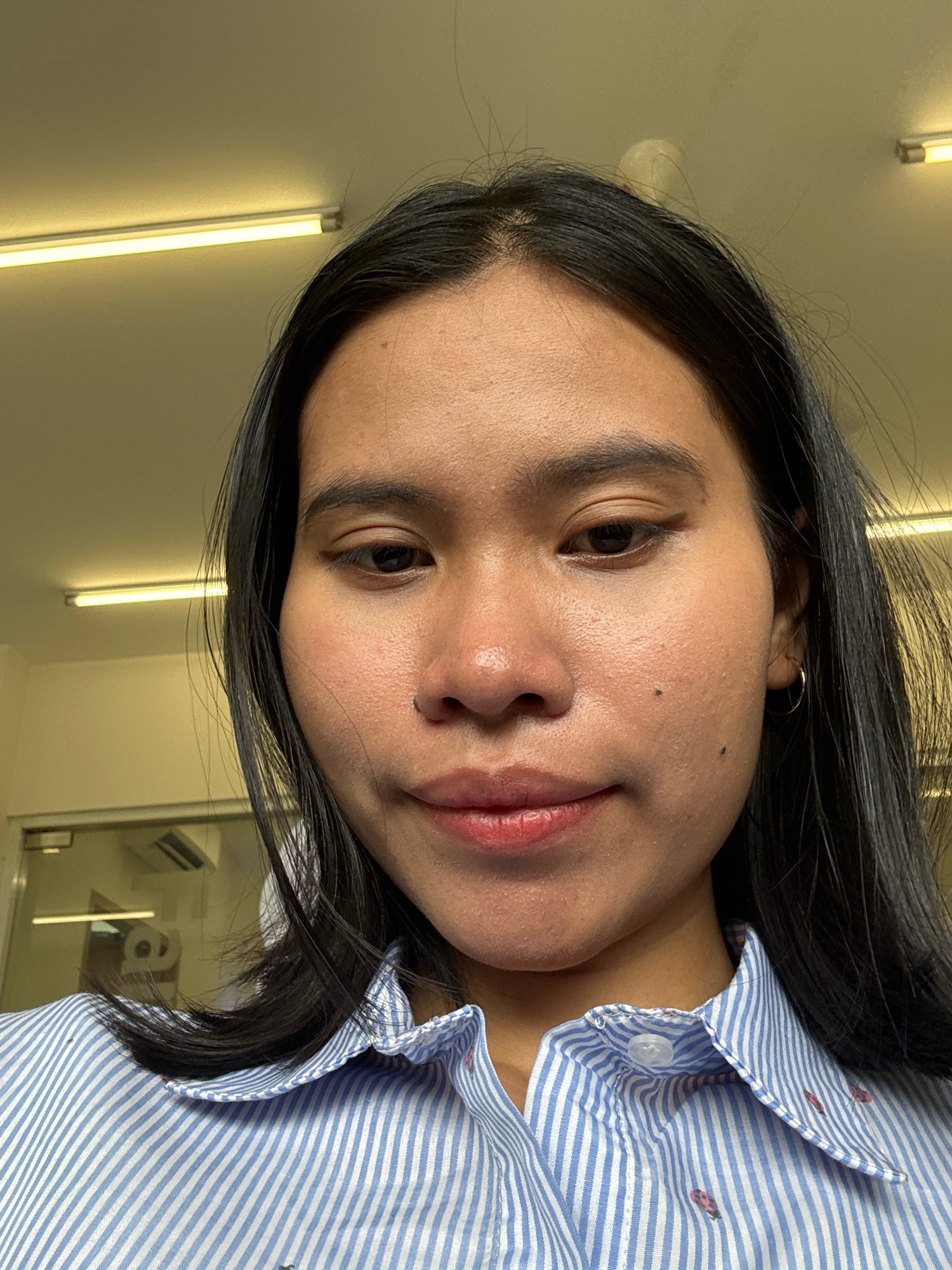 A medium-shot, indoor, selfie shows a young woman with dark hair and light brown skin wearing a blue and white striped collared shirt. Her head is tilted down and to the left. She has a small mole on her right cheek and a nose piercing. The background shows a pale yellow ceiling with fluorescent lighting. Behind her, through a glass partition, a room with a desk and a printer is visible. Based on the context provided, this image was likely taken in East Jakarta, Indonesia.