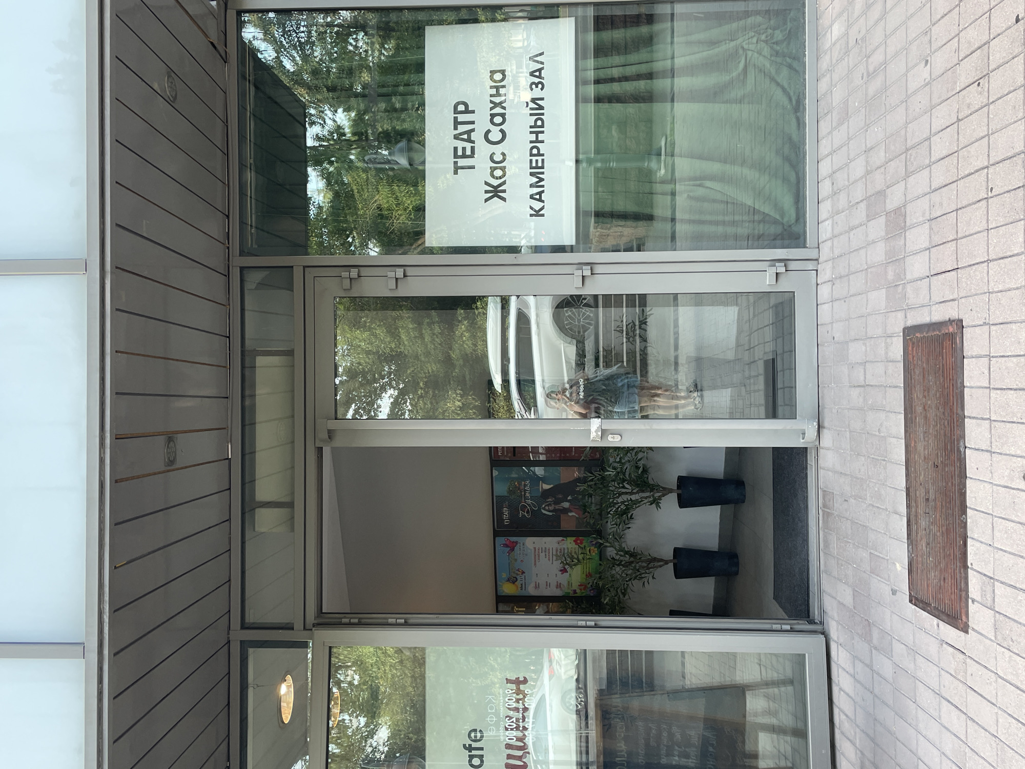 The image shows the entrance to a theater or performance space, identified by a sign that reads "ТЕАТР Жас Сахна КАМЕРНЫЙ ЗАЛ" (Theater Zhas Sakha Chamber Hall). The entrance consists of a large glass door that is partially open, revealing a glimpse of the interior. Through the glass, posters for upcoming events are visible. Two potted plants stand inside the entrance.
The exterior features a tiled walkway in front of the building, with a metal mat placed on the tiles. The building itself has a modern facade with a glass storefront and a gray overhang. In the reflection on the glass doors, a person is visible holding a phone, taking a selfie. The surrounding environment suggests an urban setting, with trees and a white car visible in the background.
Based on the presence of the "Zhas Sakha Theater" and the context of the Cyrillic script, this location is likely in Kazakhstan, and specifically Almaty, given the prominence of this theater in the city. The scene captures a typical daytime view of the theater entrance, with the signage and reflections indicating its purpose and location.