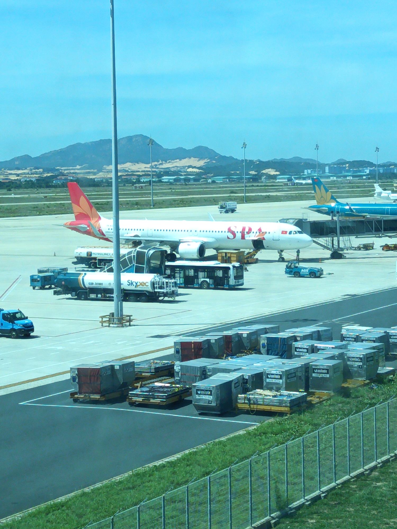 The image depicts an airport tarmac in Bắc Cam Ranh, Vietnam, under a clear blue sky. Several aircraft are visible, including a white passenger jet with red and orange markings on its tail, bearing the logo "SPA" on its fuselage. To the right, a teal airplane with a golden design on its tail is parked. Ground support equipment, such as baggage carts filled with cargo containers, a fuel truck labeled "PETROLIMEX," and a bus are positioned around the aircraft. A small blue vehicle is also present on the tarmac. In the background, hills and buildings can be seen. The foreground shows a section of a fence and grassy area.