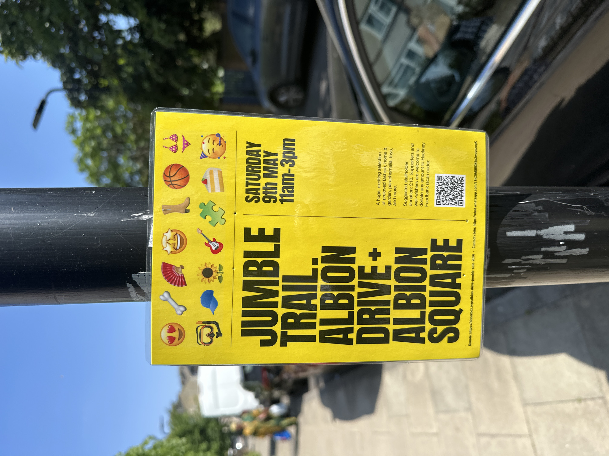 Here's a description of the image:
A yellow sign is attached to a black pole. The sign advertises a "Jumble Trail" event on Albion Drive and Albion Square, scheduled for Saturday, May 9th, from 11 am to 3 pm. The sign features a variety of emojis along the top and left side of the text. Information on the sign promotes a "huge, exciting selection of preloved fashion, home & garden, paraphernalia, toys, and more." It also lists a suggested stallholder donation of £10, encouraging supporters to donate to the Hackney Foodbank. A QR code is included, alongside links for donations and joining a WhatsApp chat.
The pole is positioned outdoors. A car, trees, buildings, and a white van are visible in the background, suggesting an urban street environment. The image appears to have been taken during the day under sunny conditions.