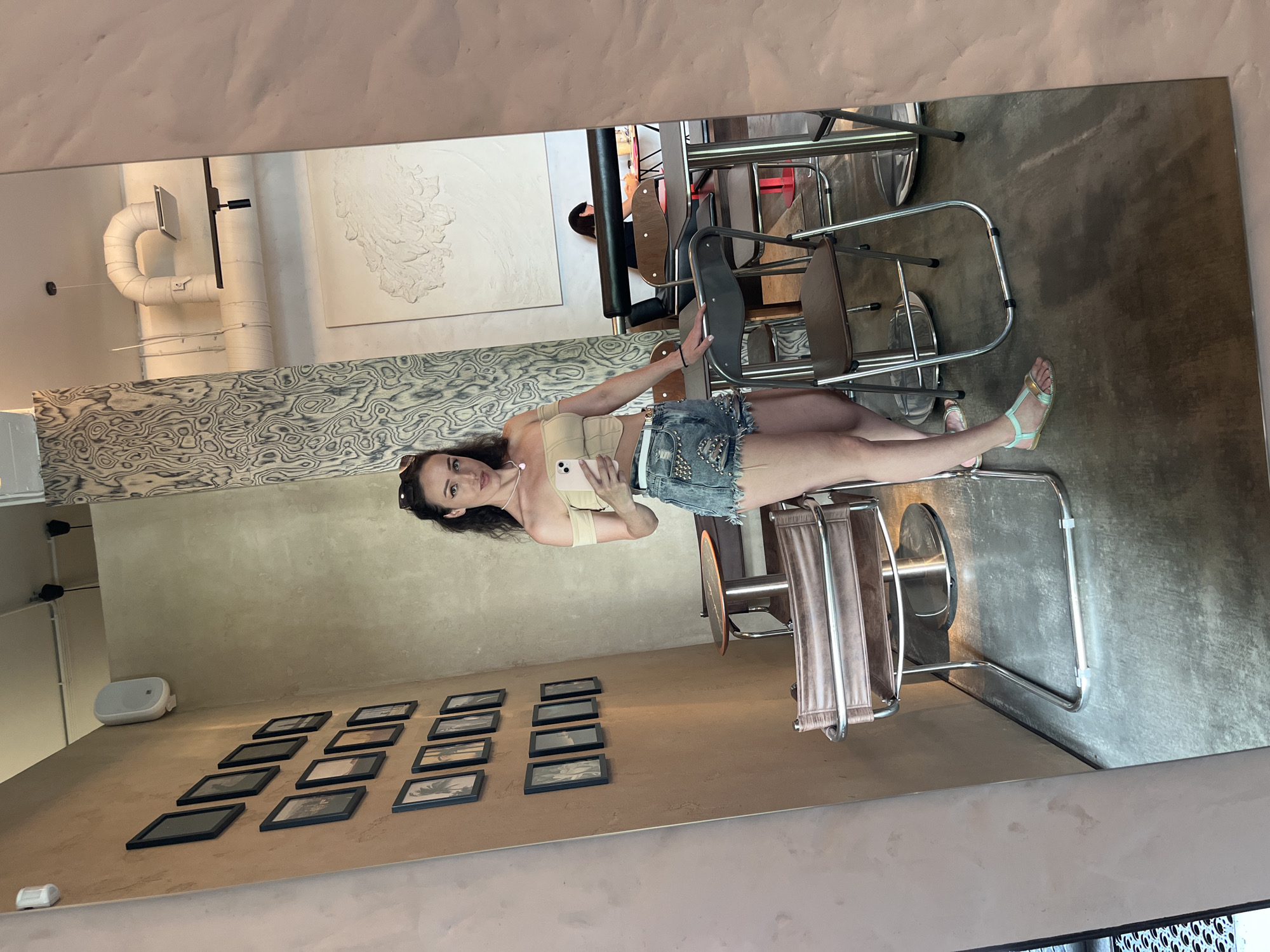 A young woman with dark hair, wearing a beige off-the-shoulder top and denim shorts, takes a mirror selfie in a cafe. She is holding a pink smartphone in her right hand and has a necklace on. She is standing next to a modern, metallic-framed chair with brown leather upholstery. The cafe has concrete floors and textured walls with framed pictures. In the reflection, other tables and chairs are visible, as well as another person sitting at a table in the background. The overall ambiance is contemporary and casual. The location is identified as a cafe in Kazan, Russia.