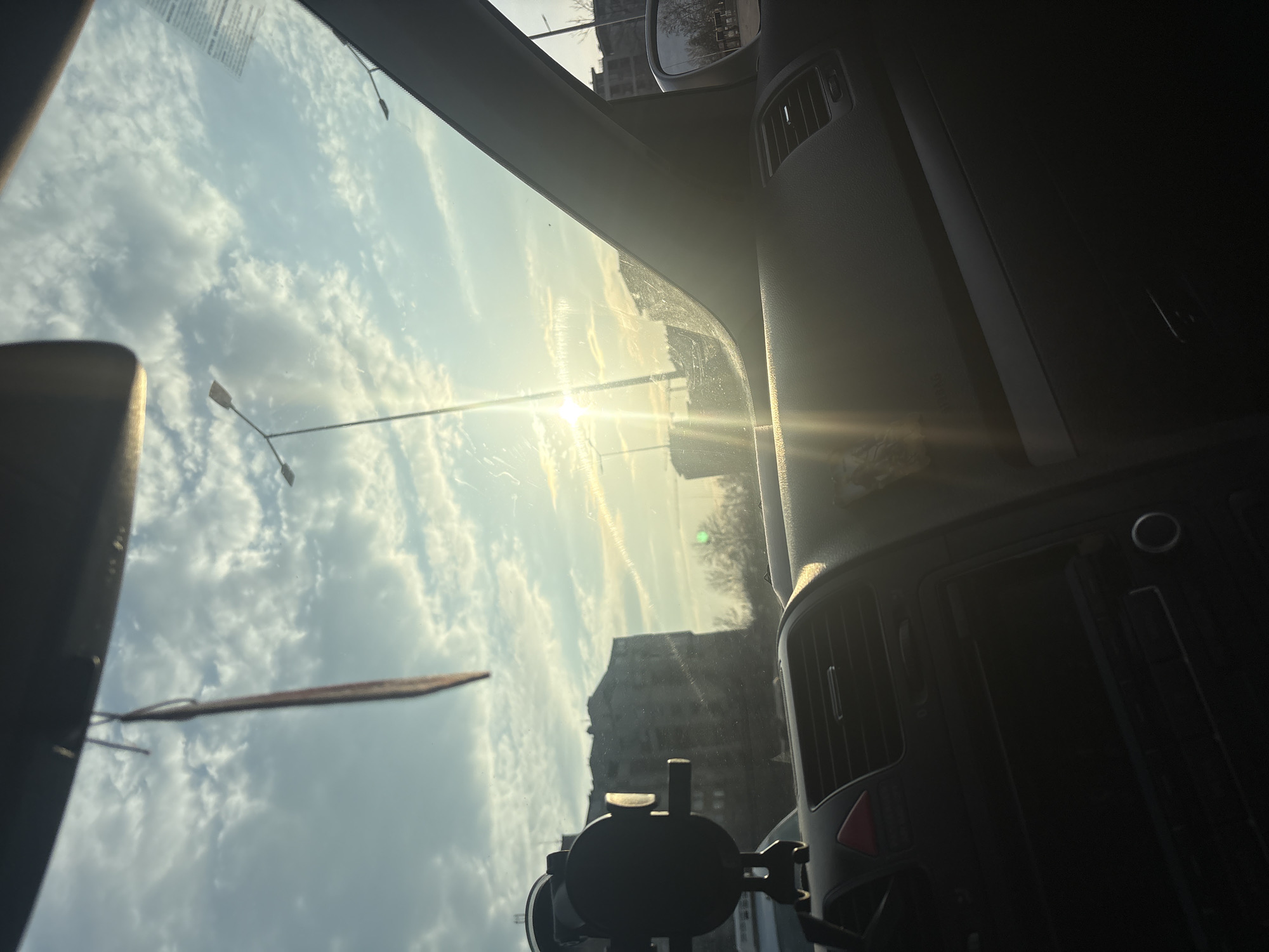 The scene is captured from within a vehicle, facing forward through the windshield. The bright sun, positioned low in the upper-right quadrant, causes significant glare and lens flare, obscuring parts of the view. Streaks are visible on the windshield surface.
The vehicle's dark-toned dashboard dominates the lower frame, displaying central air vents, a red triangular hazard light button, and an unlit infotainment system with a single rotary knob. A black suction-cup phone mount is secured to the windshield on the driver's side. A slender, dark object hangs from the interior rearview mirror area. The passenger-side A-pillar and a portion of the exterior side mirror are visible on the right.
Beyond the vehicle, a sky with scattered white clouds is visible. Multiple tall streetlights are silhouetted against the bright sky. On the left, a large, multi-story concrete apartment building with visible wear stands. Further right in the background, another structure, possibly incomplete, is discernible. Bare trees are present in the middle ground. No individuals are observed. The specific location within Plovdiv, Bulgaria, cannot be identified based on the visual information provided.