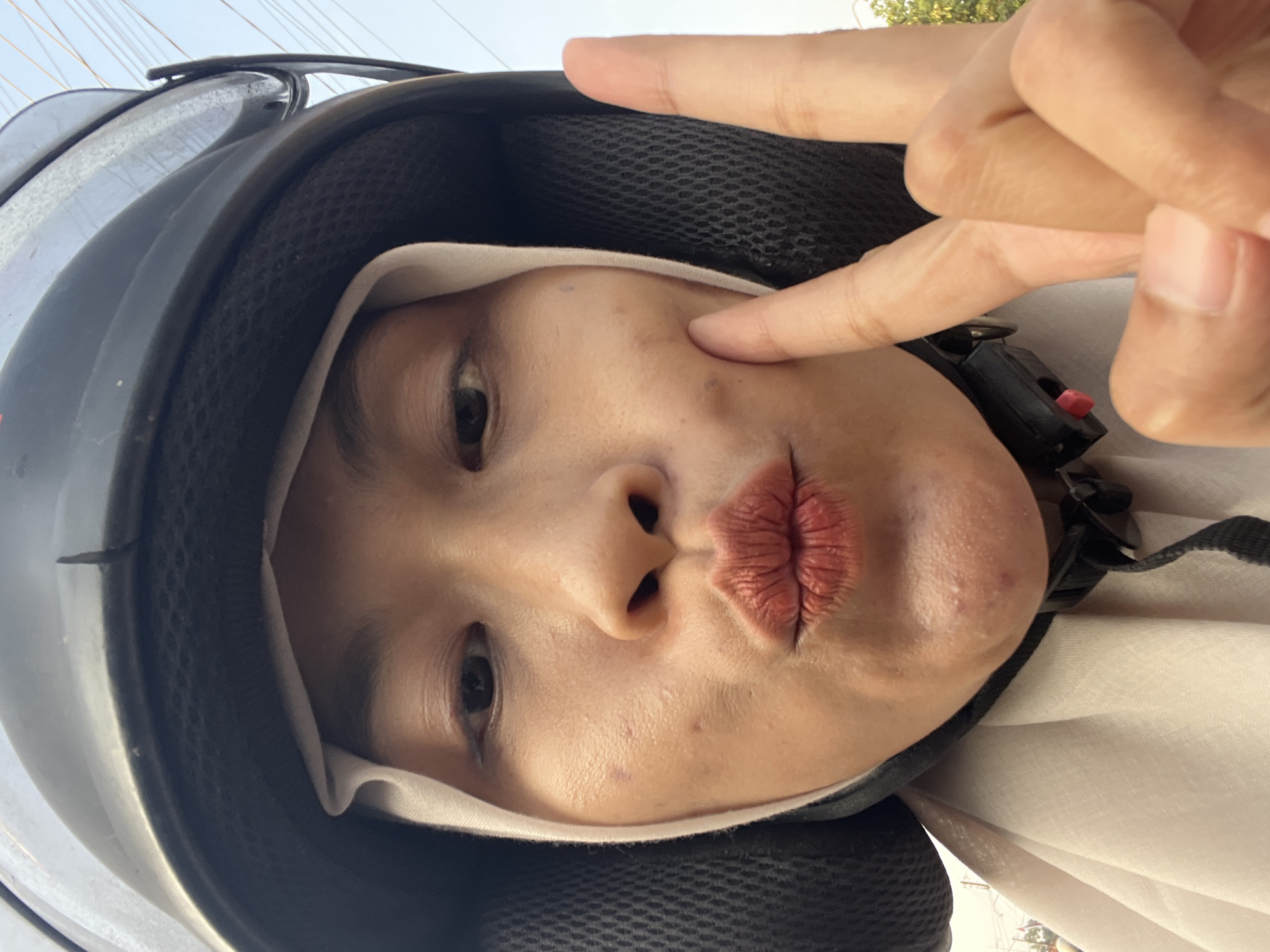 A close-up, selfie-style, eye-level shot shows a person wearing a black helmet and a light-colored hijab. The person is making a kissing face, with their lips puckered and colored with matte lipstick. Two fingers are raised in a peace sign gesture, positioned to the right of their face. The person's skin appears to have some texture, with visible blemishes and acne on the chin and cheek. The background is blurred but suggests an outdoor environment with hints of trees and possibly power lines, consistent with a setting in Kecamatan Pati, Indonesia. The lighting is natural and appears to be daytime.