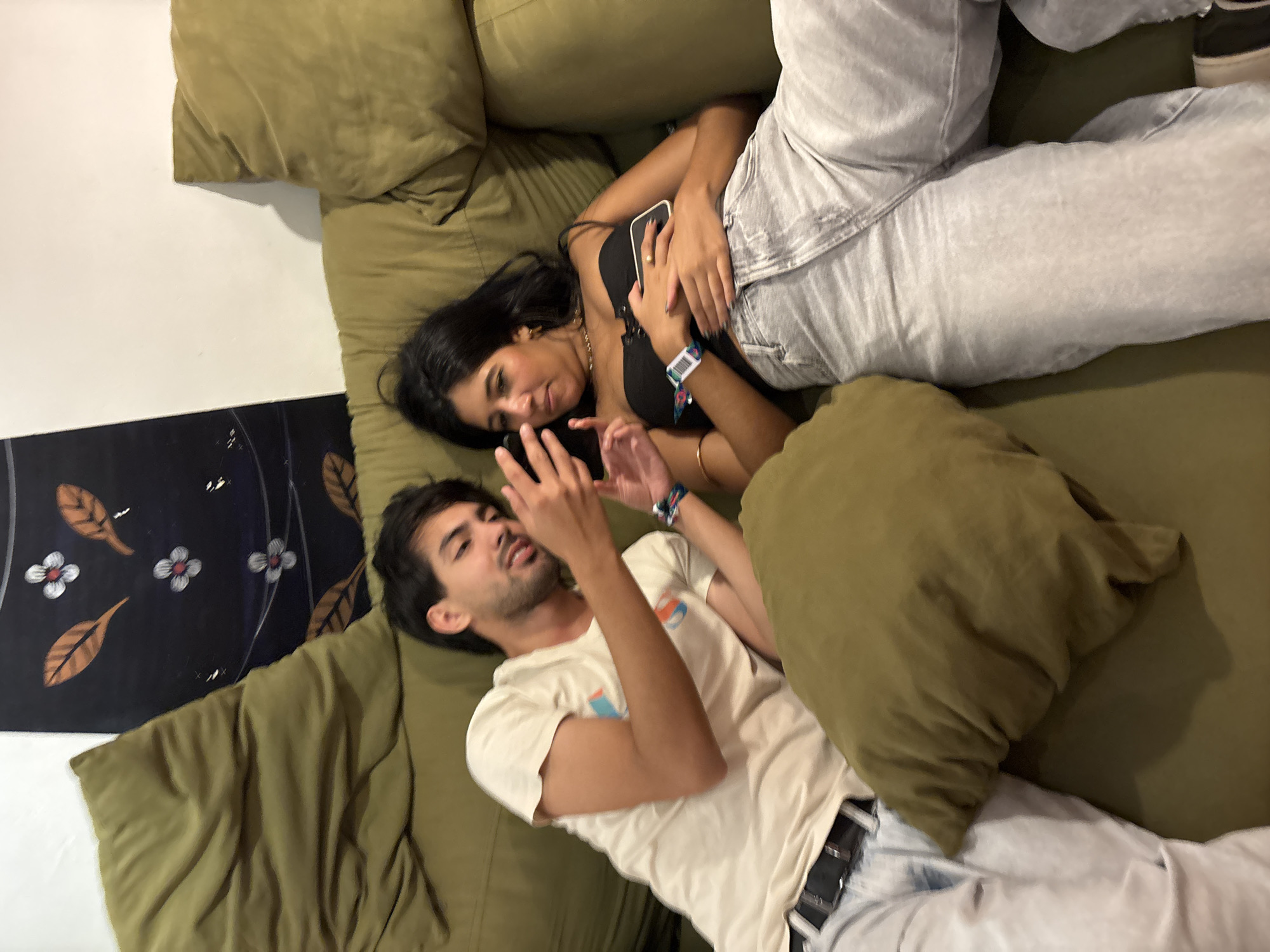 Two individuals are reclining on an olive-green couch, facing opposite directions. The woman, with dark hair, wears a black top and light grey pants. She is holding a phone. The man, with dark hair, wears a white t-shirt and light grey pants with a black belt. He is looking at his phone. A decorative floral panel is visible on the left side of the frame. Pillows, in the same olive-green hue as the couch, are scattered around. The location is possibly in an apartment or residence, in São Paulo, Brazil.