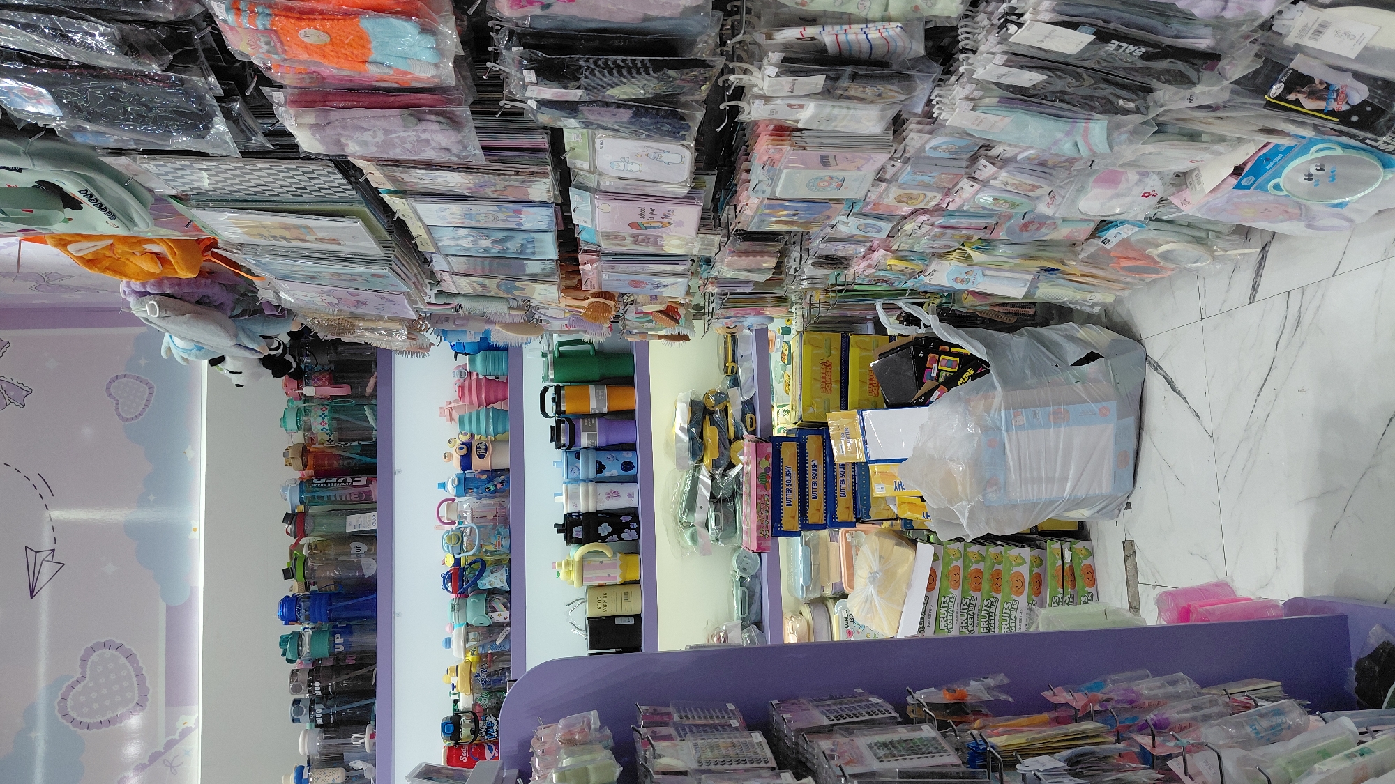 This is a well-lit, medium-angle interior shot of a retail store, likely a gift shop or a store selling children's accessories, located in Purwokerto, Indonesia. The store has a playful and colorful decor, with pastel purple shelving and walls adorned with whimsical cloud and heart motifs.
The shelves are stocked with a variety of items. On the top shelf, a row of colorful water bottles and tumblers are neatly arranged. Below them, more bottles and containers are displayed, some featuring cartoon characters. The lower shelves are filled with boxed food containers, plush toys, and smaller packaged goods.
To the right of the shelves, a wall is covered with numerous hanging items, predominantly small accessories like socks, gloves, hair accessories, and stationery, all individually packaged in clear plastic. The items are displayed on hooks, creating a dense visual effect.
In the center of the image, on the floor, a large, translucent plastic bag is filled with what appears to be more merchandise, possibly boxes or larger items. A pink plastic container is visible near the bottom left corner, and the floor is covered in white marble-patterned tiles.
There are no people visible in the frame, and the overall impression is one of a neatly organized retail environment displaying a wide array of consumer goods. The lighting is bright and even, suggesting artificial lighting common in indoor commercial spaces. The presence of specific branding or product types is not clearly discernible from this vantage point.