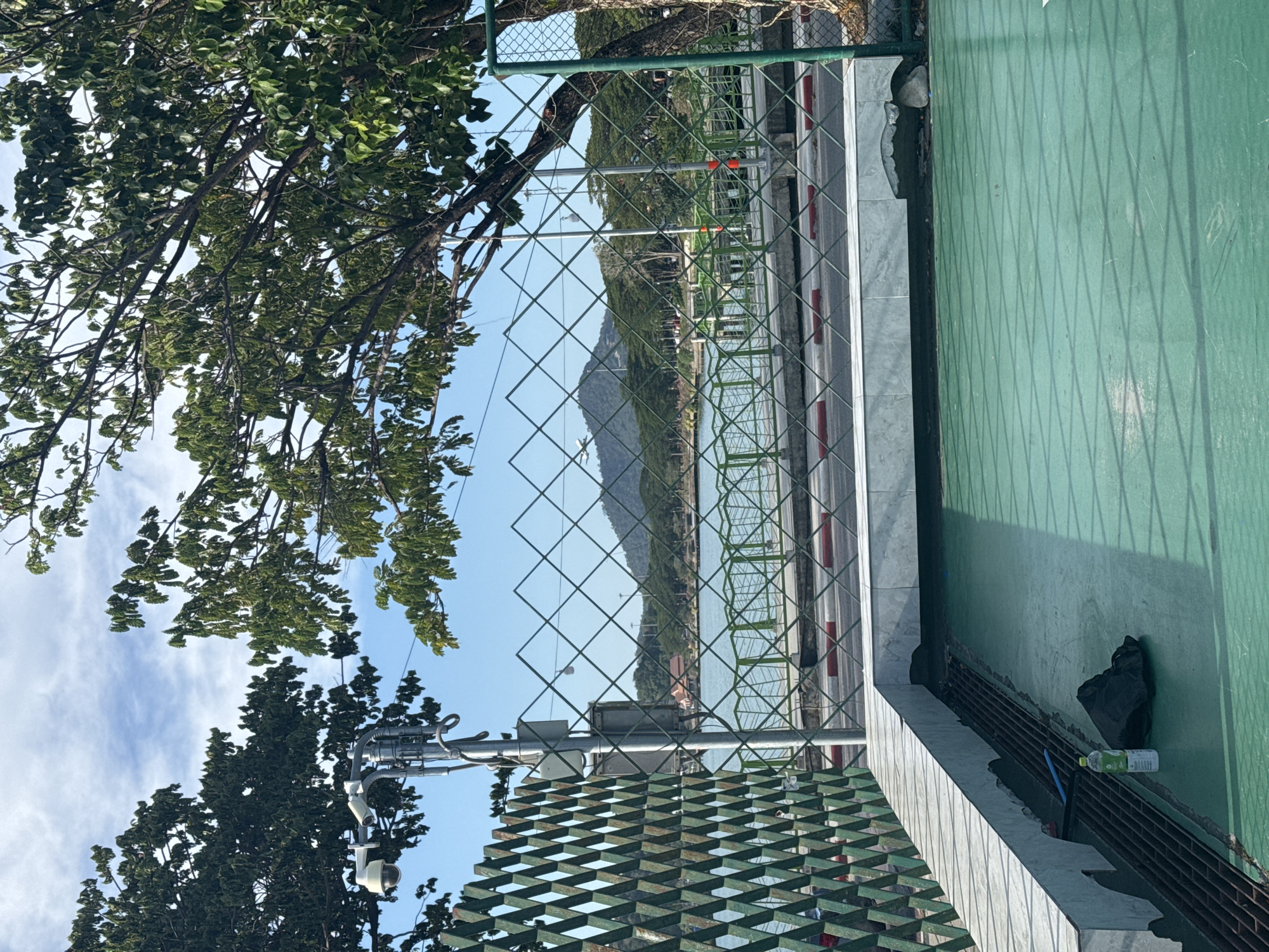 The image depicts a view through a diamond-patterned green metal fence onto a waterfront promenade on the island of Ko Samui, Thailand. In the foreground, a green, slightly worn surface, possibly a court or deck, is visible with a white marble-like structure along its edge. A partially damaged section of the marble structure reveals debris beneath. On this surface, a plastic water bottle and a black bag are placed.
Beyond the fence, a body of water is visible, likely the sea or a bay, with a promenade running along its edge. The promenade has a low green railing and a paved walkway with red and white curbing. Trees line the far side of the promenade, and in the distance, a lush green hillside with a prominent peak is seen under a bright blue sky with scattered clouds. A faint white object, possibly an airplane, is visible ascending near the distant hillside.
To the left, a utility pole with two security cameras and electrical equipment is mounted, partially obscured by the foliage of a large tree. The overall scene is one of a public outdoor space, potentially a park or recreational area, overlooking a scenic coastal landscape. The lighting suggests it is daytime, with sunlight casting shadows through the fence. There are no people visible in the image.