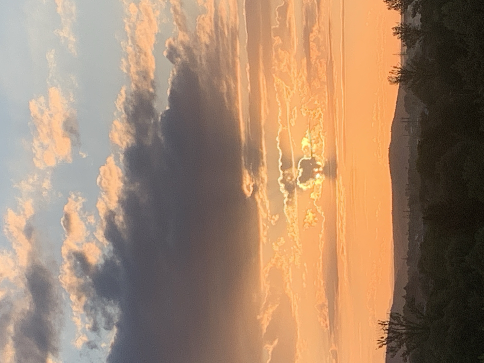 The image shows a sunset over a city, possibly Kolomyia, Ukraine. The sky is filled with dramatic clouds, illuminated by the setting sun. The sun itself is partially obscured by clouds, creating a glowing effect. In the foreground, there is a silhouette of trees and buildings, indicating an urban or suburban landscape. The overall mood is serene and peaceful, with the warm colors of the sunset creating a sense of tranquility.