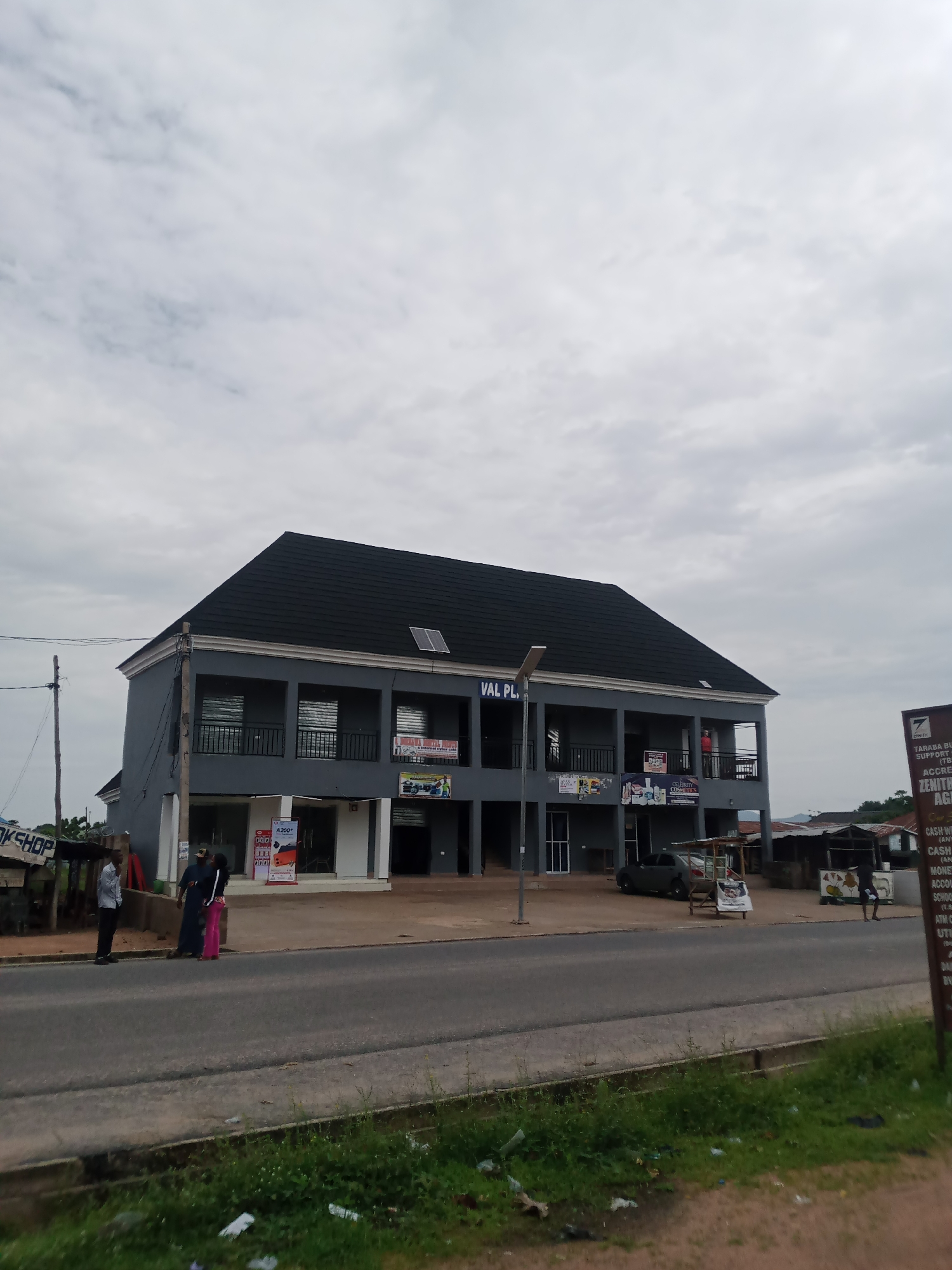 A medium shot captures a two-story gray building with a black tiled roof in Jalingo, Nigeria. The building features a prominent "VAL PL" sign on its upper level. Below the sign, several windows with blinds are visible, and a balcony extends across the front of the building. The ground floor has large glass windows and doors, indicating shopfronts. Various signs are displayed on the building, including "CELEBRITY COSMETICS" and advertisements for services like "CASH WITH ATM".
In front of the building, a paved road runs horizontally across the lower portion of the image. Several people are present: three individuals stand on the sidewalk near the left side of the building, two of whom appear to be conversing; another person stands on the balcony of the building. A car is parked in front of the shops, and various stalls and smaller structures are visible further down the street. The sky is overcast, with a cloudy, gray atmosphere. To the right, a tall sign for "ZENITH BANK" is prominently displayed. The overall scene depicts a commercial area in an urban setting.