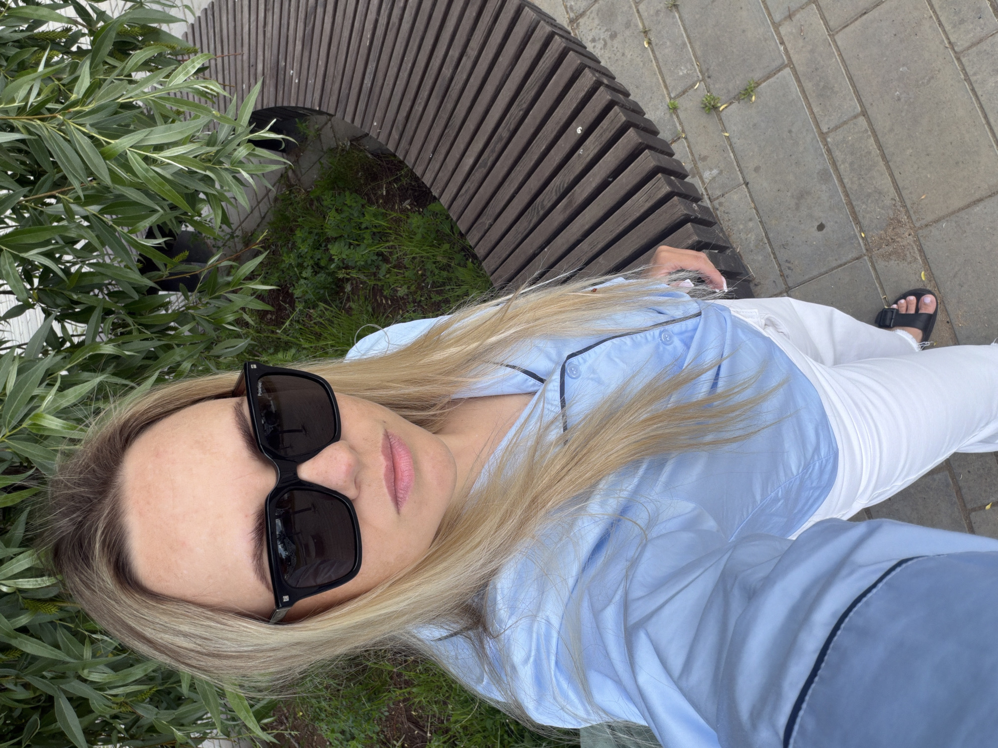 The image captures a blonde-haired individual, likely female, in a high-angle shot looking directly at the camera. She is wearing large black rectangular sunglasses, a light blue long-sleeved button-down shirt with black piping along the placket and cuffs, and white trousers. A black sandal with a wide strap is visible on her right foot.
The scene is set outdoors, featuring a curved public bench made of dark brown wooden slats. The ground is partly paved with grey rectangular pavers and partly covered with green grass and leafy foliage, possibly from a willow tree. The lighting is soft and even, suggesting it is daytime, likely under overcast conditions or during early morning/late afternoon.
A notable detail is the word "POLAROID" visible on the right arm of the sunglasses. The person's expression is neutral, with her eyes obscured by the sunglasses. The overall setting suggests a casual outdoor location, such as a park or public walkway, in Votkinsk, Russia.