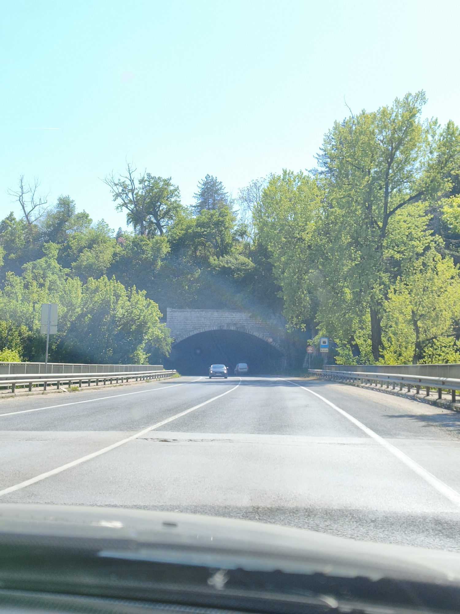 The image shows a view from inside a car traveling along a road towards a tunnel in Veliko Tarnovo, Bulgaria. The road has two lanes separated by a solid white line. Guardrails line both sides of the road. A vehicle is visible exiting the tunnel. The tunnel is surrounded by green trees and foliage. The sky is clear and blue. The dashboard and windshield of the car are visible in the foreground.