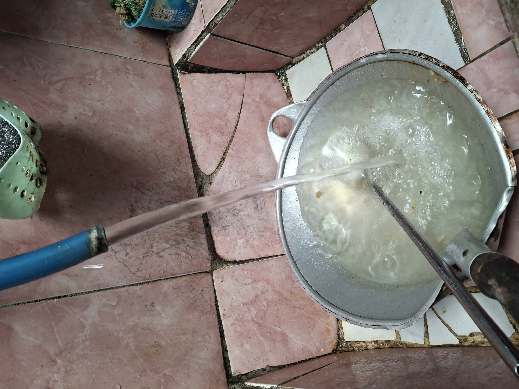 In Lumajang, Indonesia, a scene unfolds in what appears to be an outdoor sink area or kitchen workspace. Pink and white tiles, some cracked and stained, form the walls and floor, with dark grout lines. A blue hose, slightly worn at the end, directs a stream of water into a large, metallic wok. The wok, also showing signs of use and rust, is filled with murky water and some food debris. A metal utensil, possibly a stirrer, rests inside the wok. To the right, a light blue plastic container holds green items, and a green plastic basket with holes is visible above and to the left, containing what looks like steel wool scrubbers. The overall impression is one of a utilitarian space in use for cleaning or food preparation.
