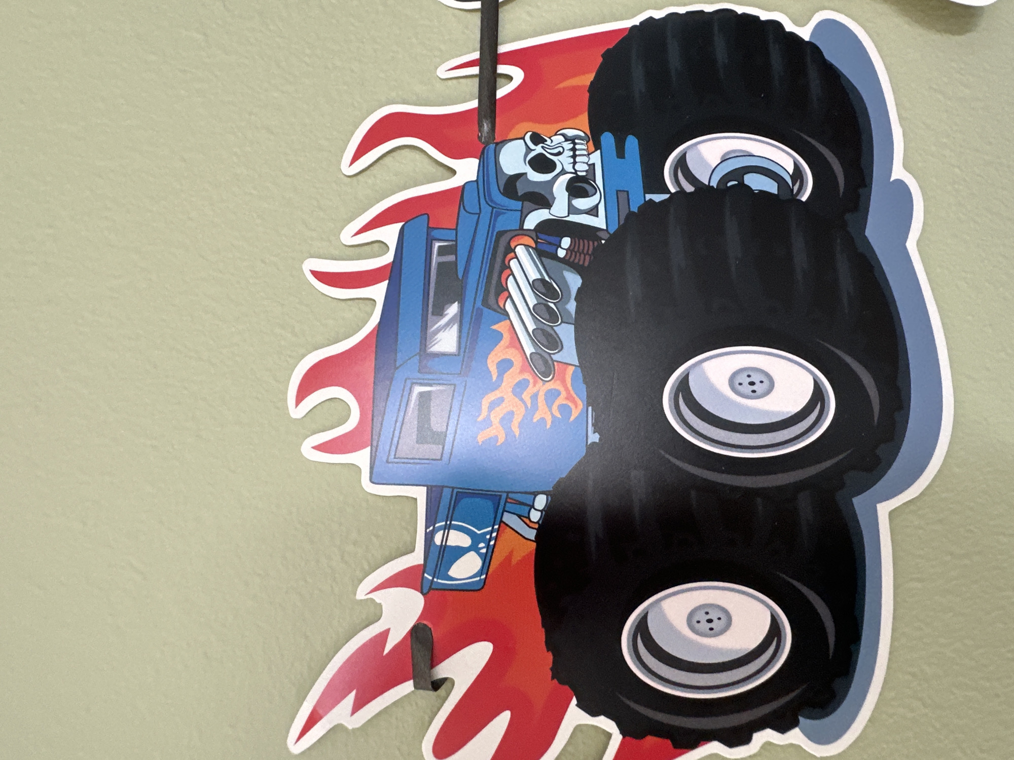 This is a close-up, vertical shot of a cut-out decoration, likely a sticker or decal, adhered to a light green wall. The decoration depicts a stylized monster truck with a skeletal driver. The truck itself is primarily blue, with flames rendered in orange and red adorning its side. Multiple large, black tires with silver rims are prominently featured, suggesting the truck is in motion or posed in an aggressive stance. The skeletal driver, depicted with a white skull and dark eye sockets, appears to be gripping the steering wheel. The decoration also includes elements of red flames erupting from the truck's chassis and exhaust pipes. A thin, dark rod extends vertically from the top of the decoration, possibly a hanger or part of the mounting mechanism. The overall aesthetic is cartoony and energetic, possibly intended for a child's room or a party. There is no visible text in the image.
