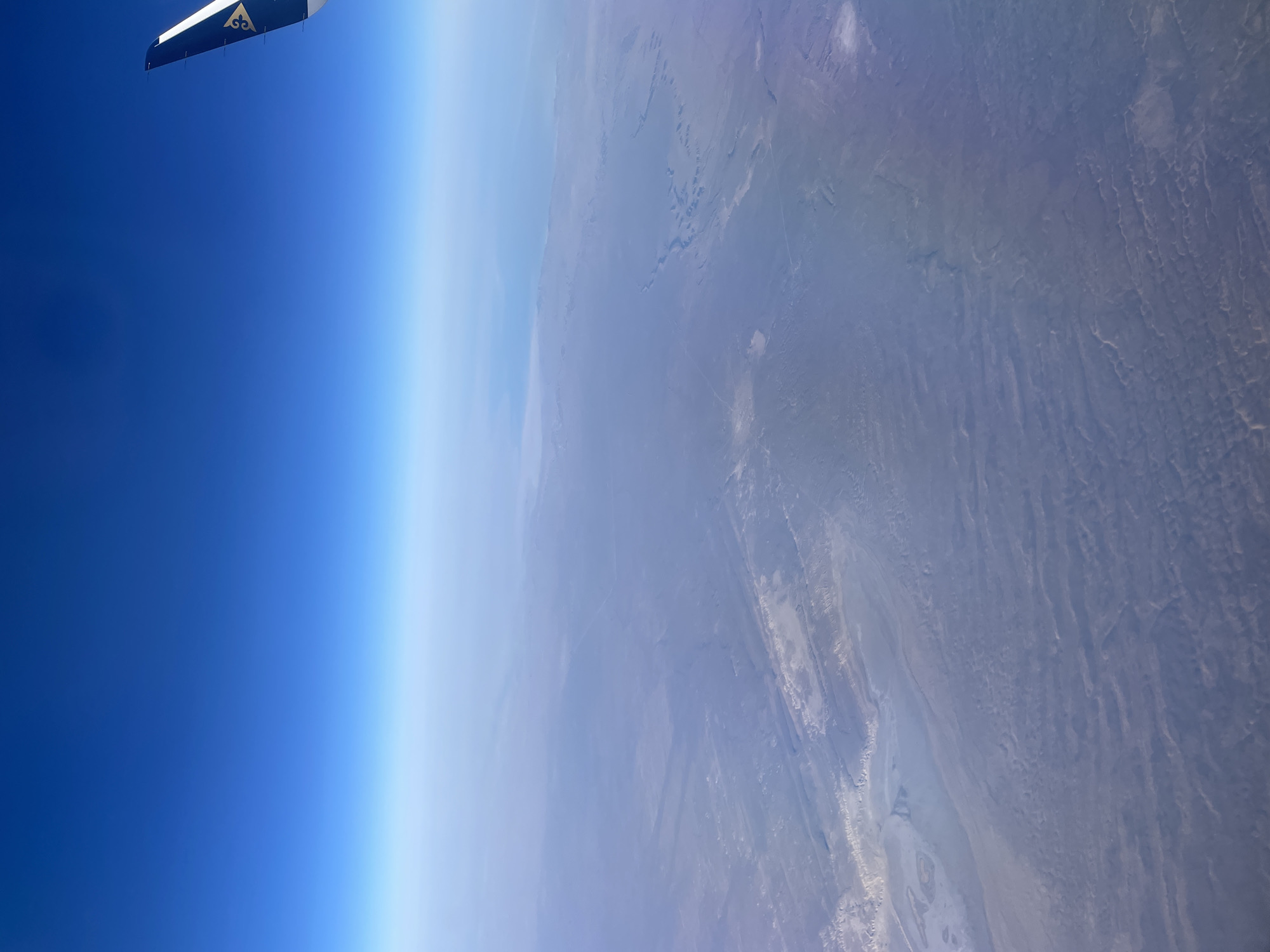 The image, taken from an airplane window, depicts a vast, hazy landscape from high altitude. The sky above transitions from a deep, vibrant blue to a lighter, almost white hue towards the horizon. The earth below appears as a muted, textured expanse of browns and grays, characterized by subtle variations in topography, possibly indicating arid or semi-arid terrain with formations like dry riverbeds or eroded hills. A portion of an airplane wing, featuring a dark blue and white design with a gold emblem, is visible in the upper right corner, indicating the viewpoint is from an aircraft in flight. The haze suggests atmospheric conditions that obscure finer details of the landscape. There are no discernible people, animals, or man-made structures identifiable in the terrestrial view.