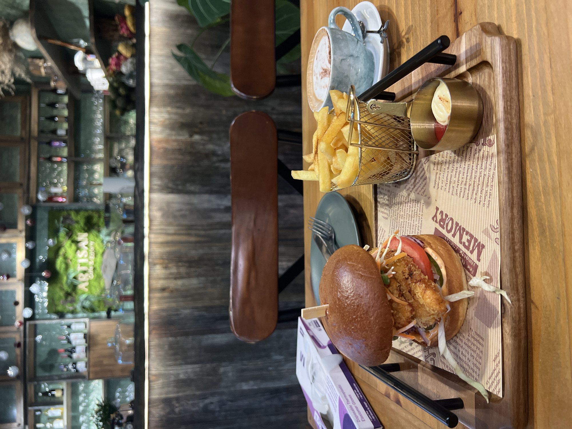 The image depicts a meal served on a wooden table in a restaurant or cafe setting. The main focus is on a wooden serving board holding a burger, fries served in a small metal basket, and a small bowl of dipping sauce, likely mayonnaise and ketchup. A blue cup of coffee is also placed on the board. A fork is on the table near the burger. A box of disposable thermoplastic elastomer gloves is on the table near the burger.
The table is accompanied by dark brown chairs. In the background, a bar area is visible, featuring shelves with wine bottles and a decorative wall arrangement including the words "ARTISAN ECO".
The picture was taken from an elevated perspective, possibly from the viewpoint of a seated customer. The photo suggests a dining establishment within a city. The location is Nha Trang Ward, Vietnam.