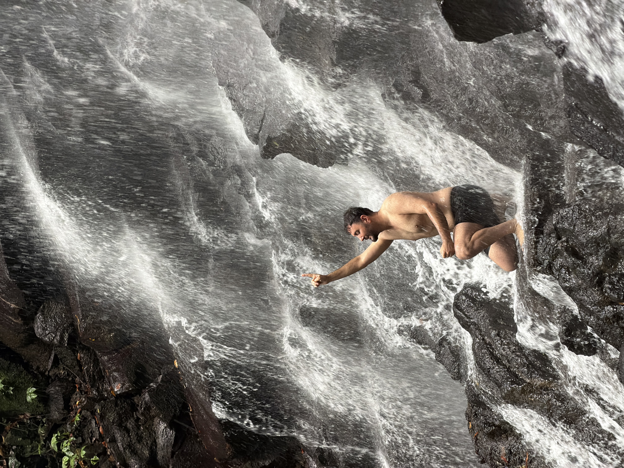 A shirtless man is kneeling on dark, wet rocks in front of a waterfall. He is wearing black swim trunks and has his left arm raised with his index finger pointing upwards. The waterfall cascades down a rocky cliff face, with white water creating a misty effect. The surrounding environment is lush and green, suggesting a natural, tropical setting. The image appears to be taken in the city of Gianyar, Indonesia, a region known for its numerous waterfalls.