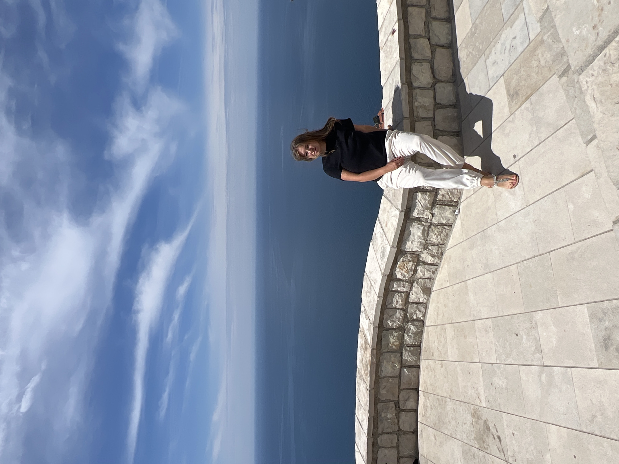 A woman is sitting on a stone ledge, likely on the coast near Sveti Stefan, Montenegro. The sky is clear and blue with wispy clouds. The ocean stretches to the horizon. The woman is wearing a black shirt and white pants, and she is facing the camera. She has long brown hair. The stone ledge curves and has a rough, textured edge. The stone ground is made of rectangular slabs.