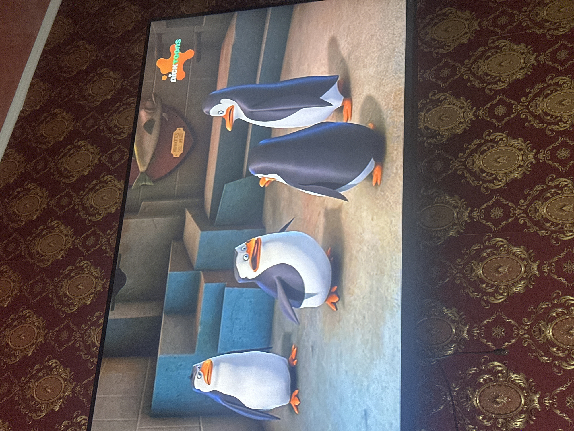 A television displays an episode of "Penguins of Madagascar." Four penguins are shown in the scene. Behind them are steps and an ornamental fish. The TV is on a patterned wallpaper.