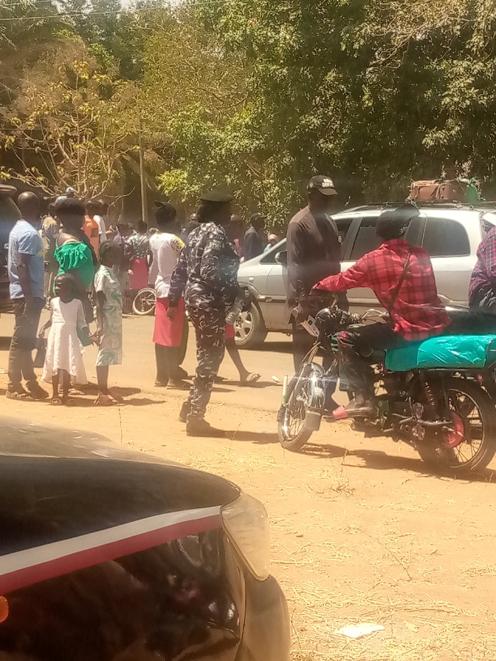The image depicts an outdoor scene in Kagoma, Nigeria, under bright daylight on an unpaved, light brown surface. Approximately twenty individuals are gathered, comprising adults and children. In the left-center, a person in a dark camouflage uniform with a dark cap stands facing left, positioned near a group of civilians. This group includes a child in a white dress and another child in a light patterned outfit. A small white child's bicycle lies on its side on the ground near these civilians. To the right, a male individual wearing a red and black plaid shirt, black trousers, and pink sandals is seated on a black motorcycle with chrome accents, facing right, and wearing a black helmet. Behind the motorcycle, a white SUV, possibly an Opel Zafira, is parked with luggage secured on its roof rack. A man in a dark shirt and a black baseball cap stands near the front of the white SUV, facing the motorcycle rider. Dense green foliage and trees form the background. A partial view of a black vehicle is visible in the immediate foreground, showing its front passenger side headlight and a red and white stripe along the hood.