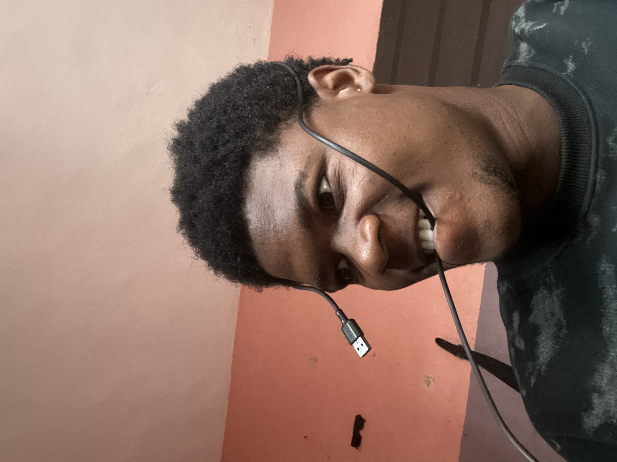 In Ihiagwa, Nigeria, a young Black person with short, dark, curly hair is captured in a close-up, head-and-shoulders shot, biting down on a black USB cable. The cable loops around their head, with the USB-A connector visible near their left ear and the cable extending towards their mouth. Their eyes are wide, looking towards the left of the frame, and their mouth is open, exposing teeth as they grip the cable. They are wearing a dark, patterned sweatshirt. The background features a coral-colored wall with some dark marks and a wooden surface to the right. The lighting is somewhat dim, with the subject illuminated from the front.
