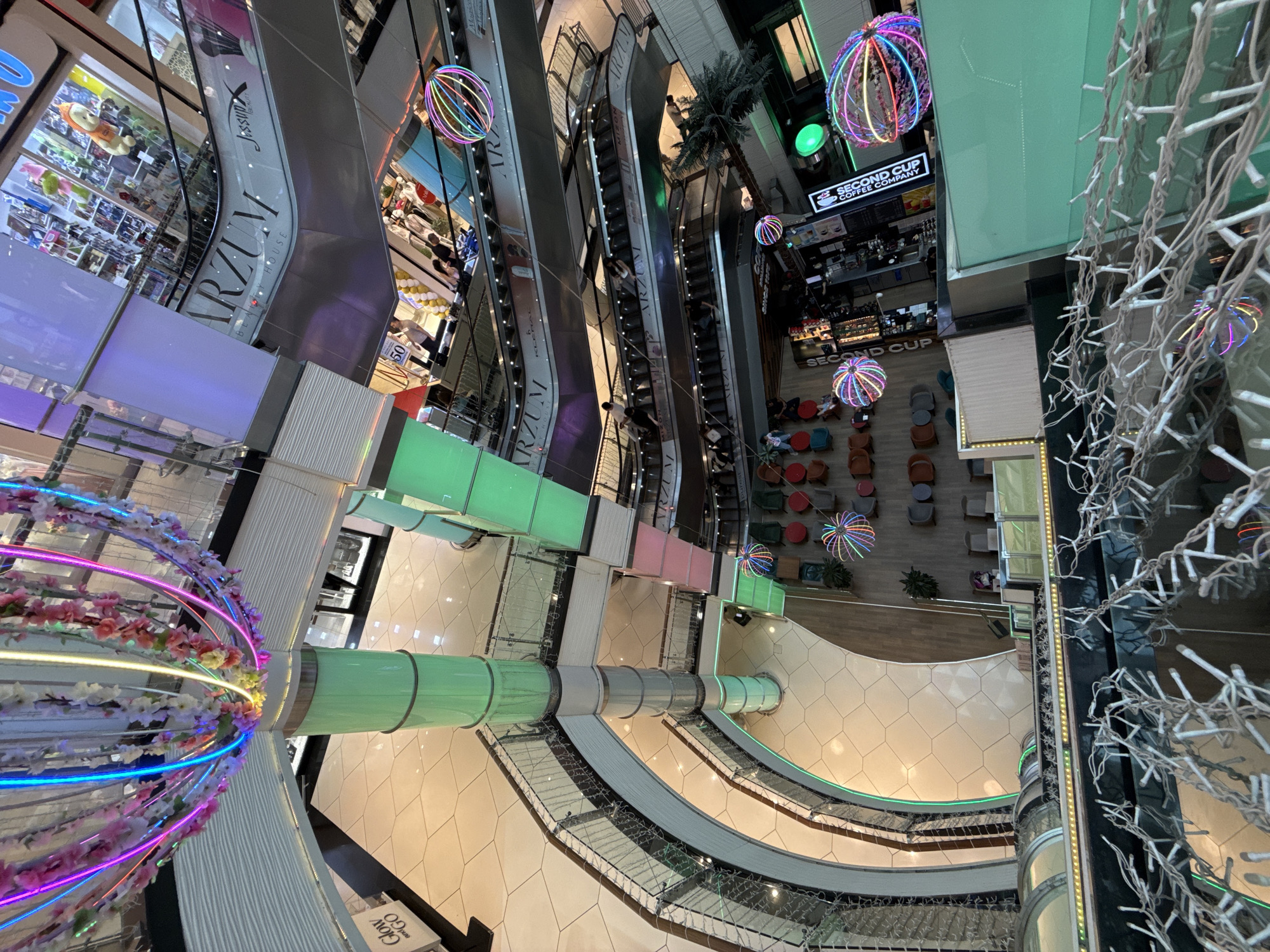 The image depicts the interior of a multi-level shopping mall in Baku, Azerbaijan, likely Port Baku Mall, given the presence of the ARZUM brand visible on escalators. The perspective is from a high vantage point, looking down into the atrium and multiple floors.
Dominating the scene are escalators that ascend through the mall, with shoppers moving on them. Various retail stores are visible on each level, some with displays of merchandise. Prominently featured is the ARZUM brand name, suggesting it is a significant tenant or the name of a department store.
Decorative elements are present throughout, including colorful LED lighting and hanging floral arrangements. One notable decoration is a large, spherical light fixture composed of colorful, crisscrossing illuminated tubes, visible on multiple levels. Green cylindrical columns, also illuminated, are integrated into the mall's architecture.
In the lower section of the image, there appears to be a seating area with tables and chairs, possibly a cafe or rest area. Signs for "SECOND CUP COFFEE COMPANY" are visible, confirming a coffee shop is present. The floor is tiled, and some areas have wooden flooring. A palm tree is visible on one of the lower levels.
The overall impression is a modern, well-lit, and bustling commercial space decorated for an event or general aesthetic enhancement, with a focus on vibrant and colorful lighting.