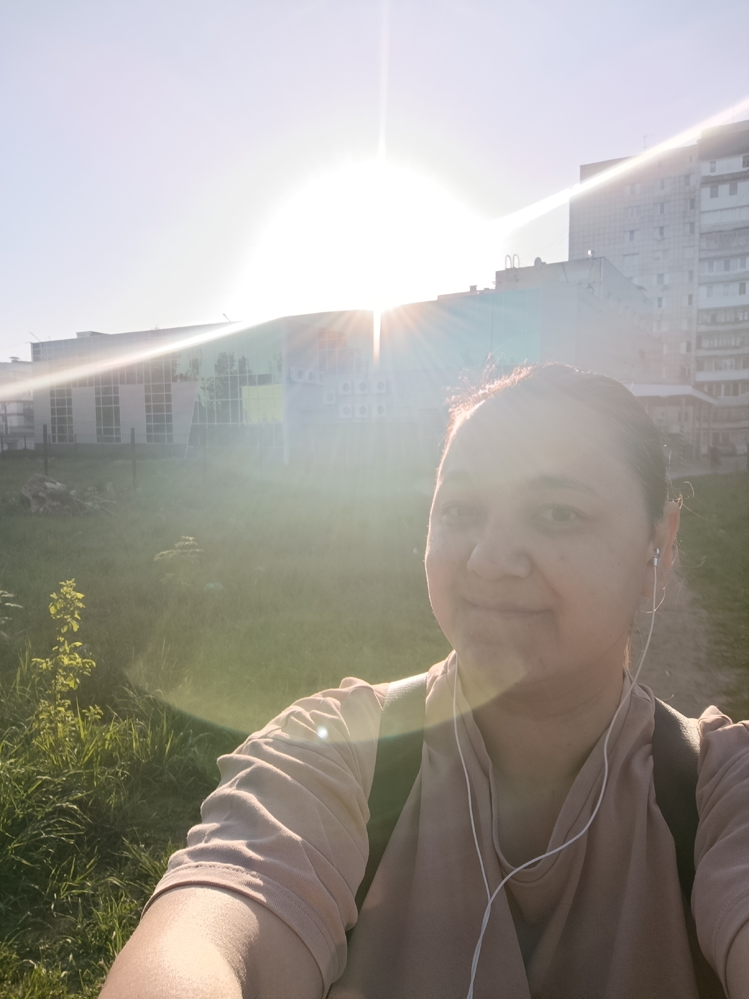 A selfie taken outdoors in the city of Chaikovsky, Russia. The subject, a woman with dark hair pulled back, wears a light brown top, a dark backpack, and white earbuds. She faces the camera with a slight smile. In the background, the sun shines brightly, obscuring some details. A multi-story building is visible to the right, and a lower structure with a glass facade can be seen to the left. Green grass and foliage fill the foreground, suggesting a park or green space. The image is overexposed due to the sun's glare.