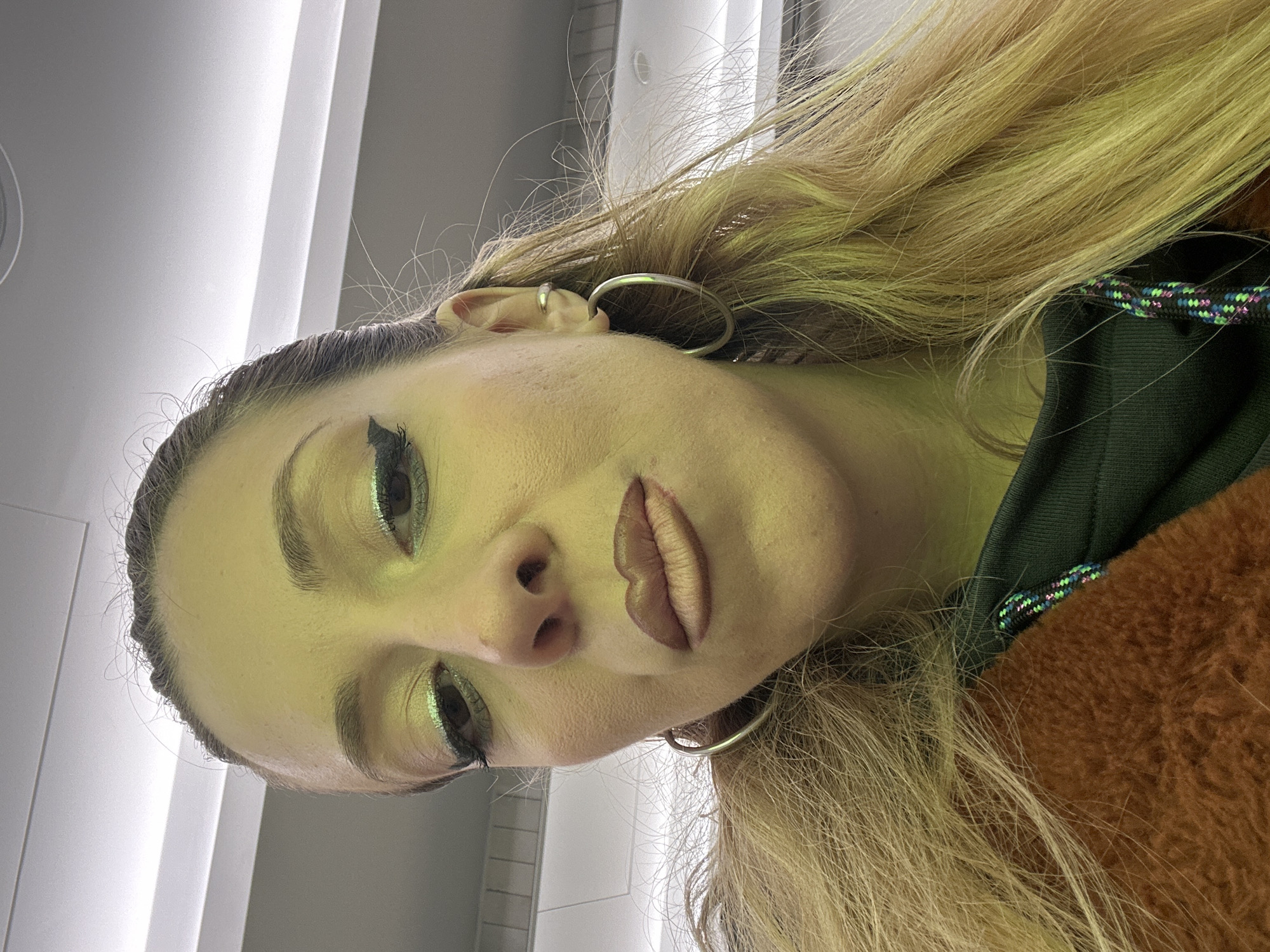 A close-up selfie of a woman with blonde hair and a dark green hoodie and brown fur jacket. She has noticeable makeup on her eyes and lips. She is wearing two silver earrings on the left ear. The background is a white wall with vertical lines. The location in the city of Dartford, United Kingdom is unknown.