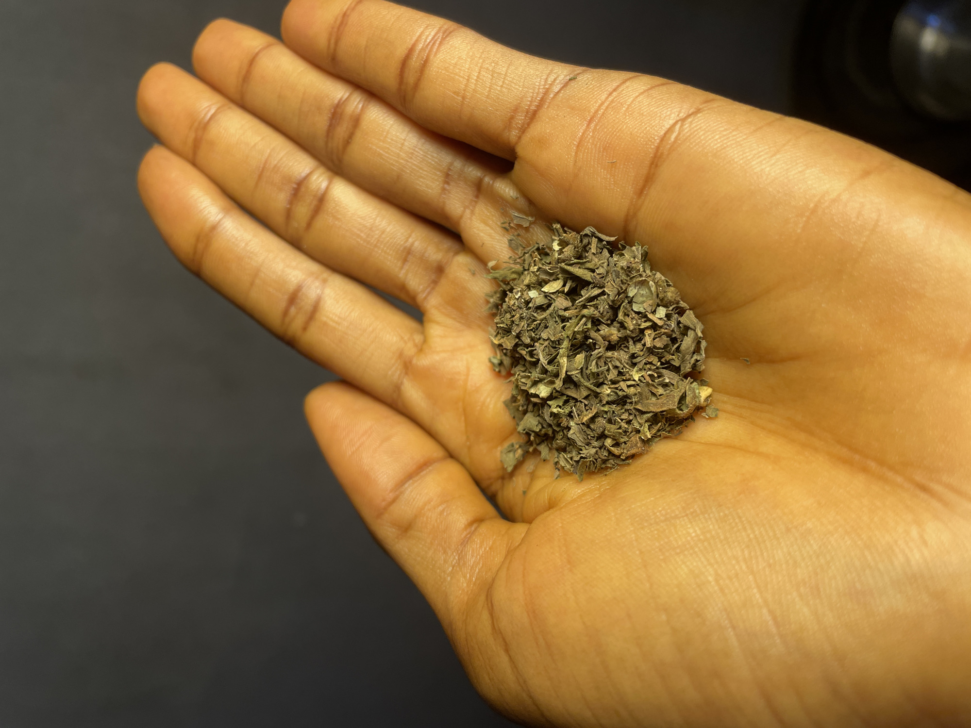 A close-up shot shows an open palm of a light-brown-skinned person, holding a small pile of dried, chopped herbs in the center. The background is dark gray, and a blurred, circular object is visible in the top right corner. No specific details suggest the location is in Nkpor, Nigeria. No other people, subjects, objects, actions, or interactions are visible.