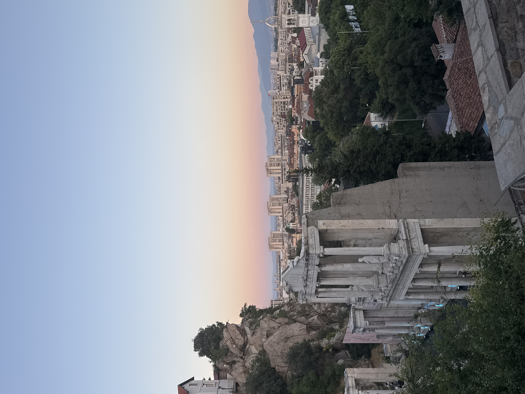 The image captures a view of the Roman Theatre of Philippopolis in Plovdiv, Bulgaria, at dusk. The ancient stone amphitheater, with its colonnaded facade and statues, is prominently featured in the foreground, partially obscured by lush green trees. In the background, the cityscape of Plovdiv unfolds, characterized by a mix of modern buildings and older structures, including a church with a distinctive tower. The sky transitions from a soft blue to hues of pink and orange, indicating the sunset. The scene is serene and historic, blending ancient ruins with the contemporary urban environment of the city.