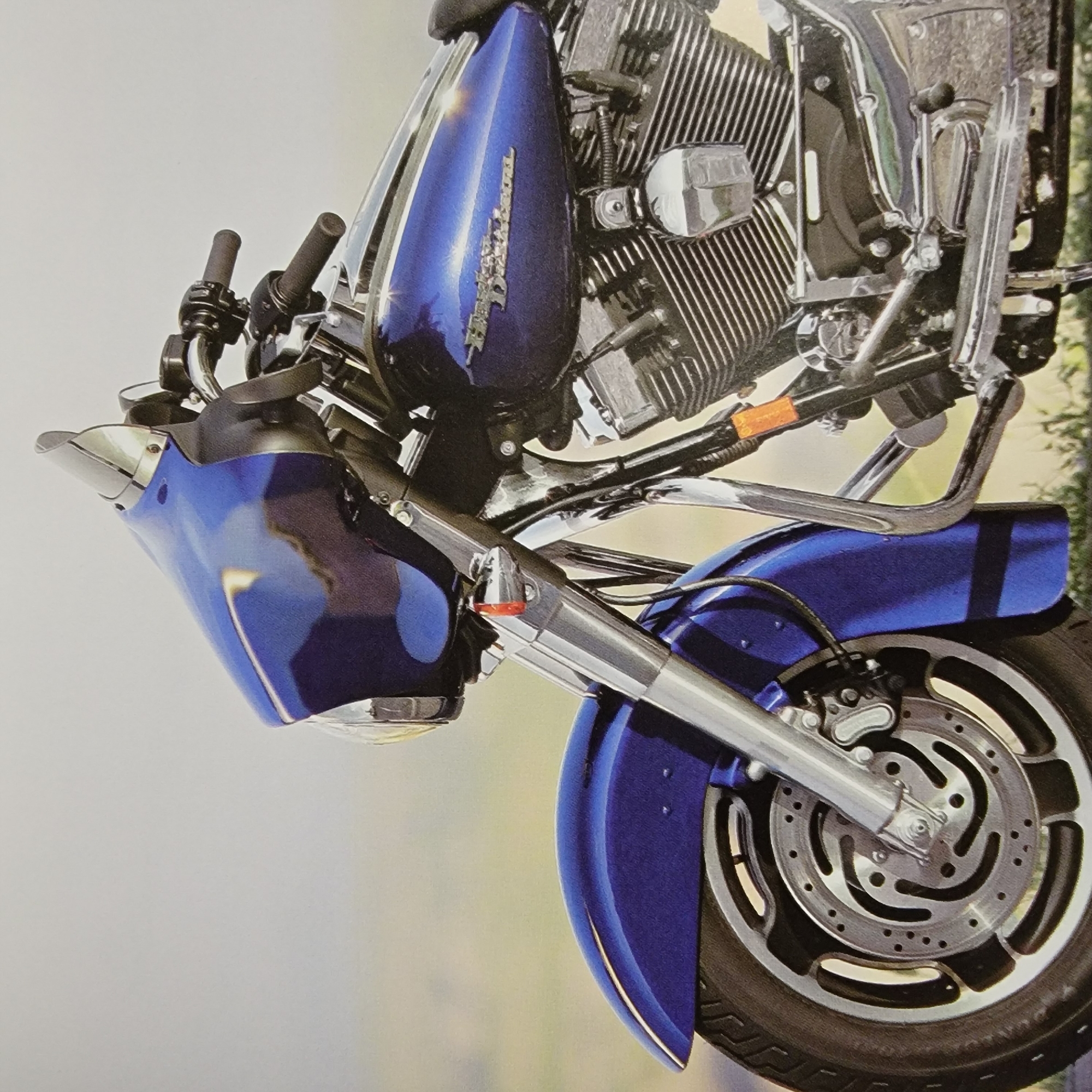 A close-up, eye-level shot shows the front portion of a dark blue motorcycle against a softly blurred light background. The motorcycle is angled, with its front wheel and fender prominently featured on the left side of the frame. The wheel is a chrome alloy with a black tire. The fender is a matching dark blue, curved and smooth. The front suspension is visible, with polished metal fork tubes.
Further into the frame, the motorcycle's fairing, also in dark blue, arches forward, revealing a clear windshield at the top. Below the fairing, the handlebars with black grips and chrome controls are visible. The motorcycle's engine, a complex assembly of black and chrome metal, is situated in the center and right of the image. The fuel tank is a deep metallic blue with a silver "Harley-Davidson" logo. Orange reflectors are visible on the motorcycle's frame. The overall impression is one of a clean, well-maintained vehicle. The background suggests an outdoor setting, possibly a field or open area, with the light suggesting a bright, possibly overcast day.