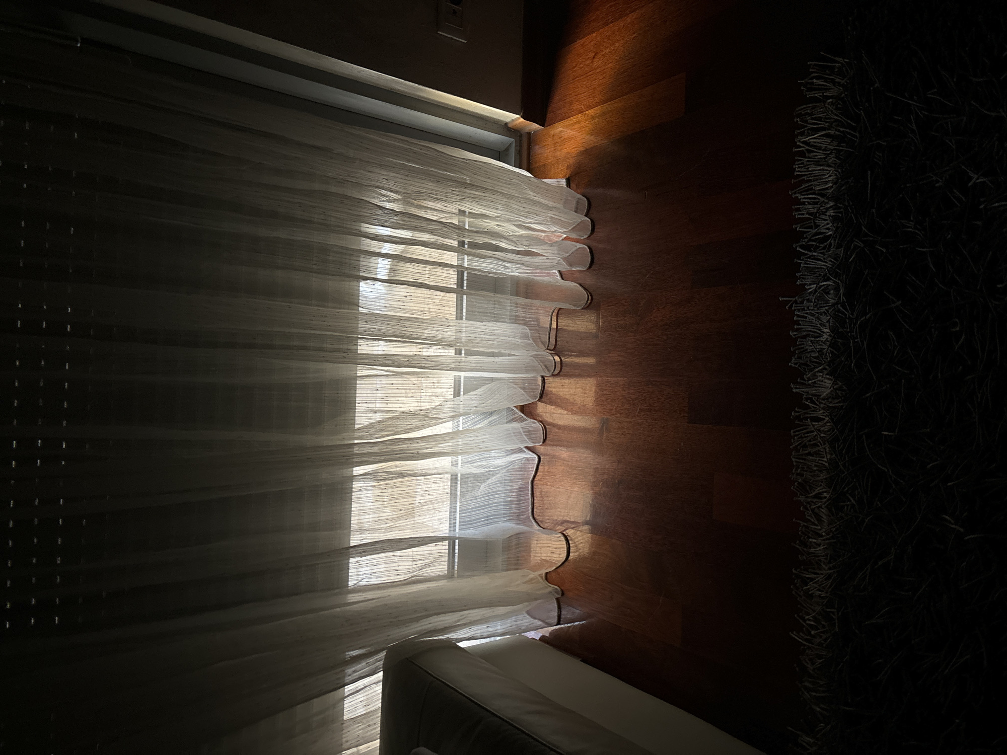 The image shows a room interior with sheer curtains covering a window. Light filters through the curtains, illuminating the wooden floor and a portion of a white sofa in the foreground. To the right of the floor is a dark, shag rug. The image includes a partial view of the walls and window frame. No people are visible. The location cannot be determined to be in Budva, Montenegro, based on the provided image.