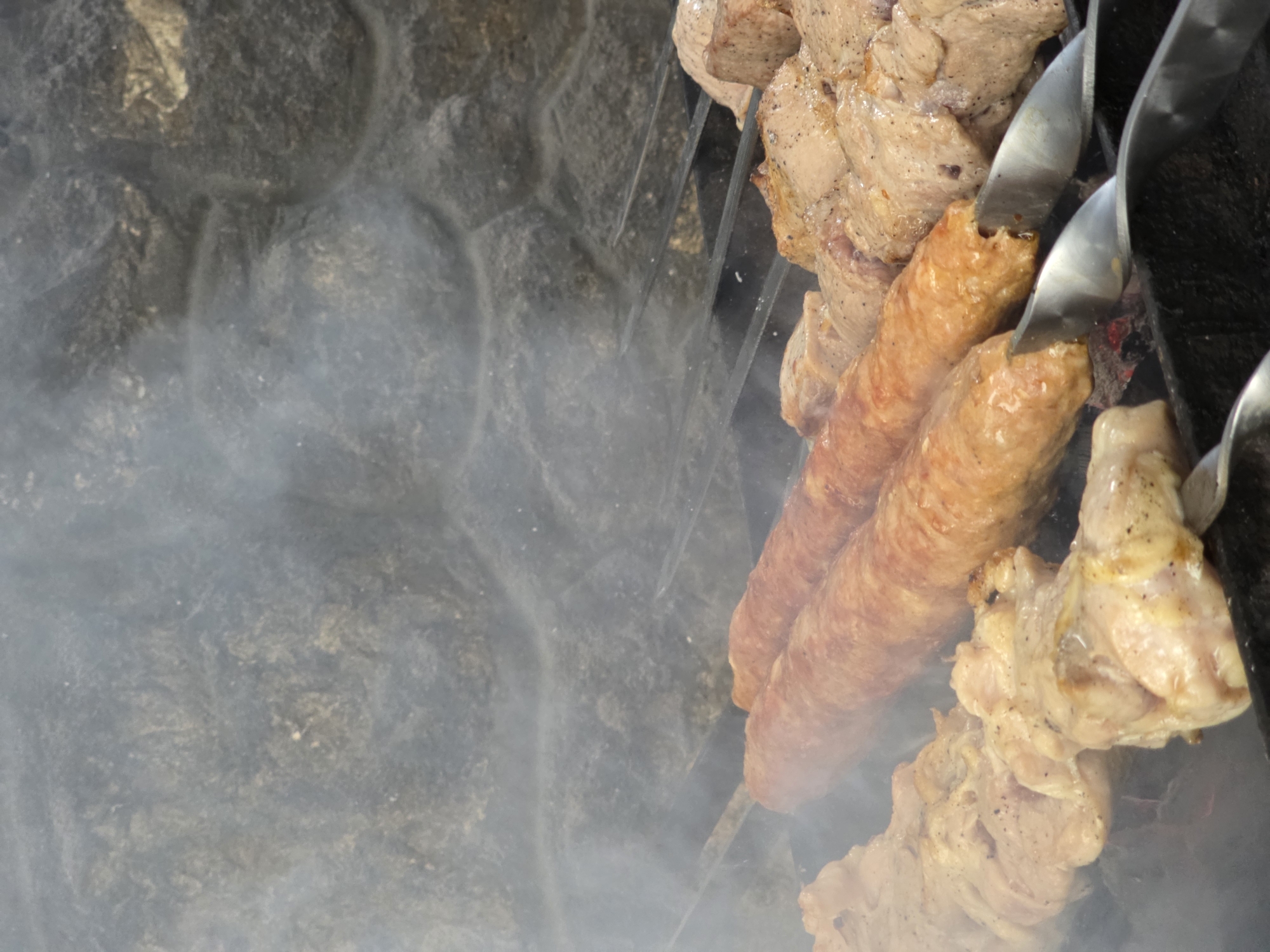 This is a close-up, eye-level shot of various types of meat being grilled on skewers over charcoal. The meat appears to be a mix of chicken pieces and long, cylindrical sausage-like kebabs. Smoke rises from the grilling meat, obscuring the background which consists of a rough, grey stone wall. The scene is likely outdoors, possibly in a backyard or park area in Pidhorodne, Ukraine, given the rustic nature of the stone and the popular barbecue activity. There are no people visible in the image. The focus is entirely on the food and the grilling process.