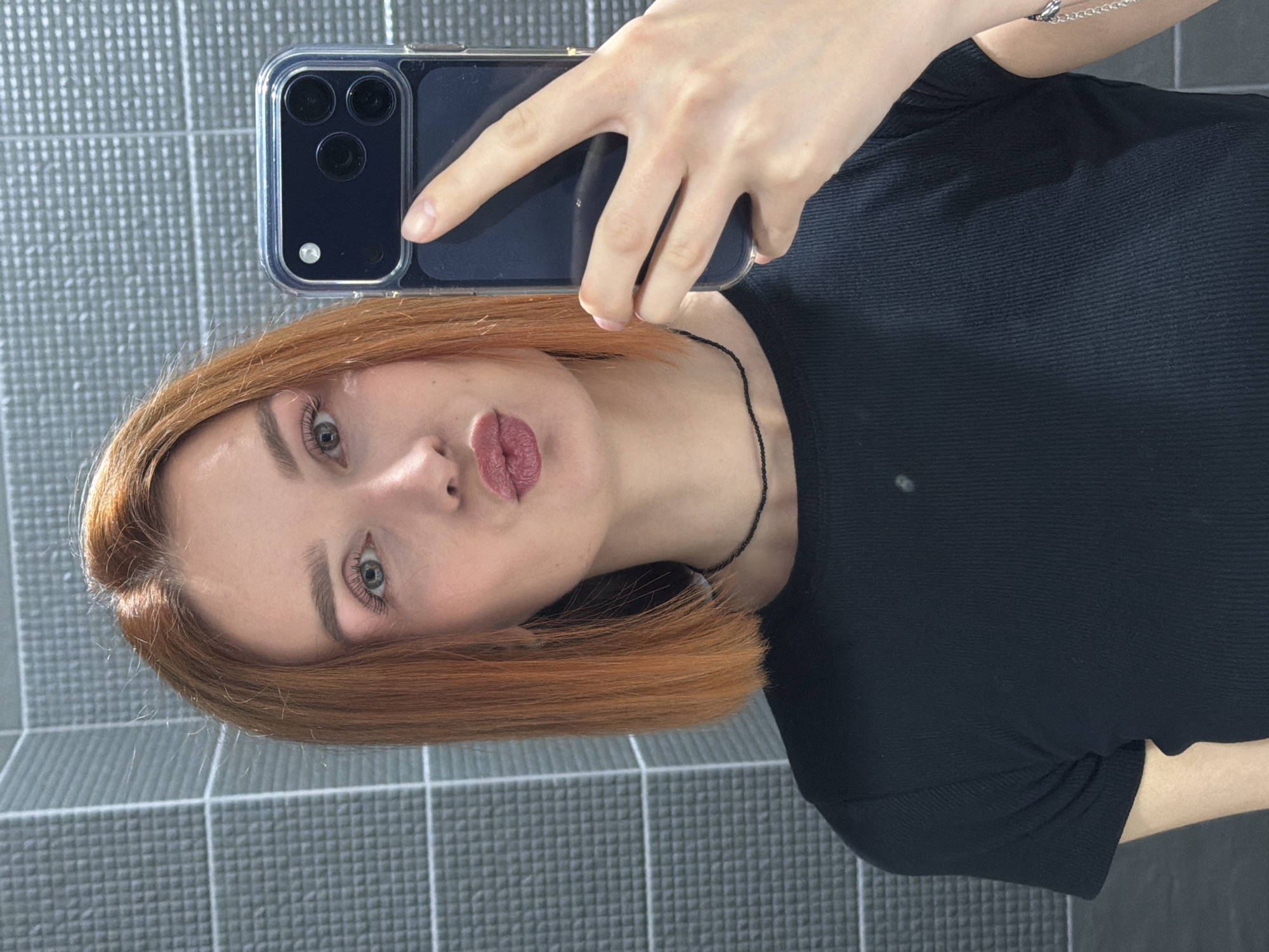 A woman with reddish-blonde hair takes a selfie using a dark-colored smartphone in a clear case. She wears a black top and a delicate silver bracelet is visible on her wrist. A thin black necklace adorns her neck. The background consists of gray tiled walls. The location is consistent with a bathroom setting, potentially in the city of Chaikovsky, Russia.