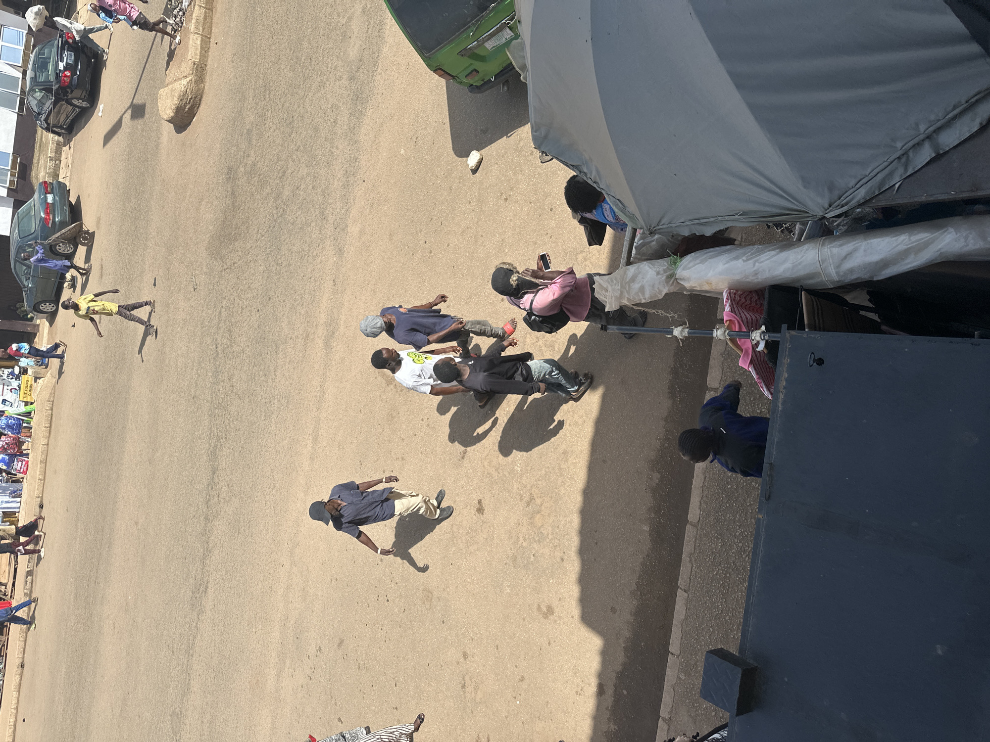 This is an aerial, eye-level, medium shot of a dusty, paved street in Bukuru, Nigeria, with a variety of pedestrians and vehicles. In the foreground, on the right, is a large, gray, collapsed tent-like structure with a rolled-up white tarp leaning against it. To the left of the tent structure, a dark blue object with a black grate is visible. Further back on the street, a group of young men are walking. One man in a dark blue shirt and a baseball cap is looking down and to his left. Another man in a white t-shirt is walking with him, and to their left, another man wears a blue t-shirt and gray pants. A man in a pink shirt and black jacket is walking away from them, looking at his phone. A green vehicle is parked on the right side of the street, behind the tent structure. Further up the street, more people are walking and there are several cars parked. One dark sedan is parked on the right, and a dark green sedan is to its left. A man in a blue shirt is pushing a wheelbarrow in front of the green sedan. A man in a yellow t-shirt is walking in the middle of the street, and another man in dark clothing is walking behind him. In the background, several stalls or shops are visible, with people browsing. The ground is a light brown, dusty color, and shadows are cast by the sun, indicating it is daytime. The overall impression is of a busy street scene in a town or city.