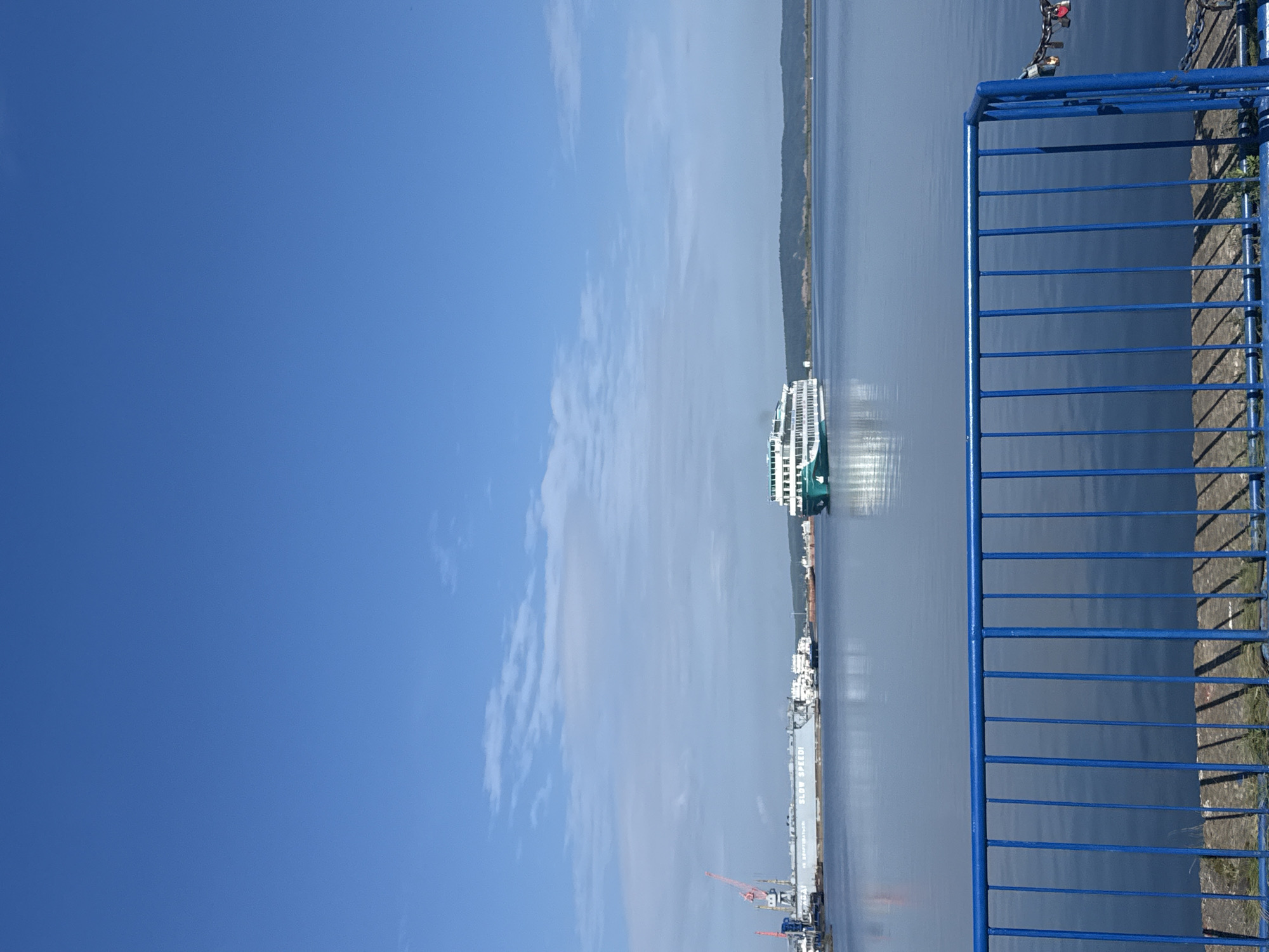 A wide-angle, eye-level shot shows a calm, blue expanse of water under a clear, bright blue sky with scattered, wispy clouds. In the foreground, a bright blue metal railing runs horizontally across the bottom of the frame. Beyond the railing, the water reflects the sky and the distant landscape.
On the left side of the image, a large industrial structure with a red crane is visible along the waterline. The structure appears to be a shipyard or port facility, with "SLOW SPEED!" written on its side. Several smaller boats are docked or moored near this facility.
Further out on the water, a white and green passenger ferry is moving from right to left across the frame. The ferry has multiple decks and appears to be a tour or transport vessel.
The background consists of a distant, dark green, tree-covered shoreline. The overall scene suggests a sunny day at a port or waterway, possibly in the city of Chaikovsky, Russia, given the context provided.