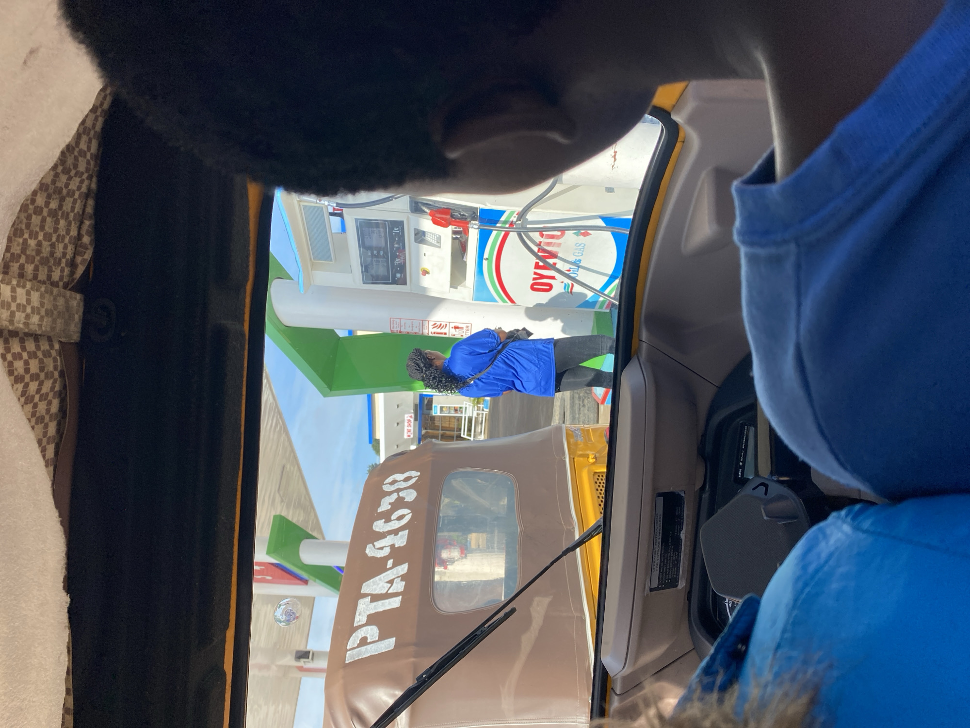This image depicts the interior of a yellow auto-rickshaw, seen from the perspective of passengers. The auto-rickshaw is parked at a gas station in Barakin Lerawi, Nigeria, as evidenced by the visible gas pumps and station signage. The license plate on the auto-rickshaw reads "PTA-4638". A person in a blue shirt is standing at a gas pump, facing away from the camera. The background shows a clear blue sky and a white gas station canopy. The foreground is dominated by the heads and shoulders of two passengers, one with dark hair and the other with lighter hair. The overall scene is casual and appears to be a typical moment during travel.