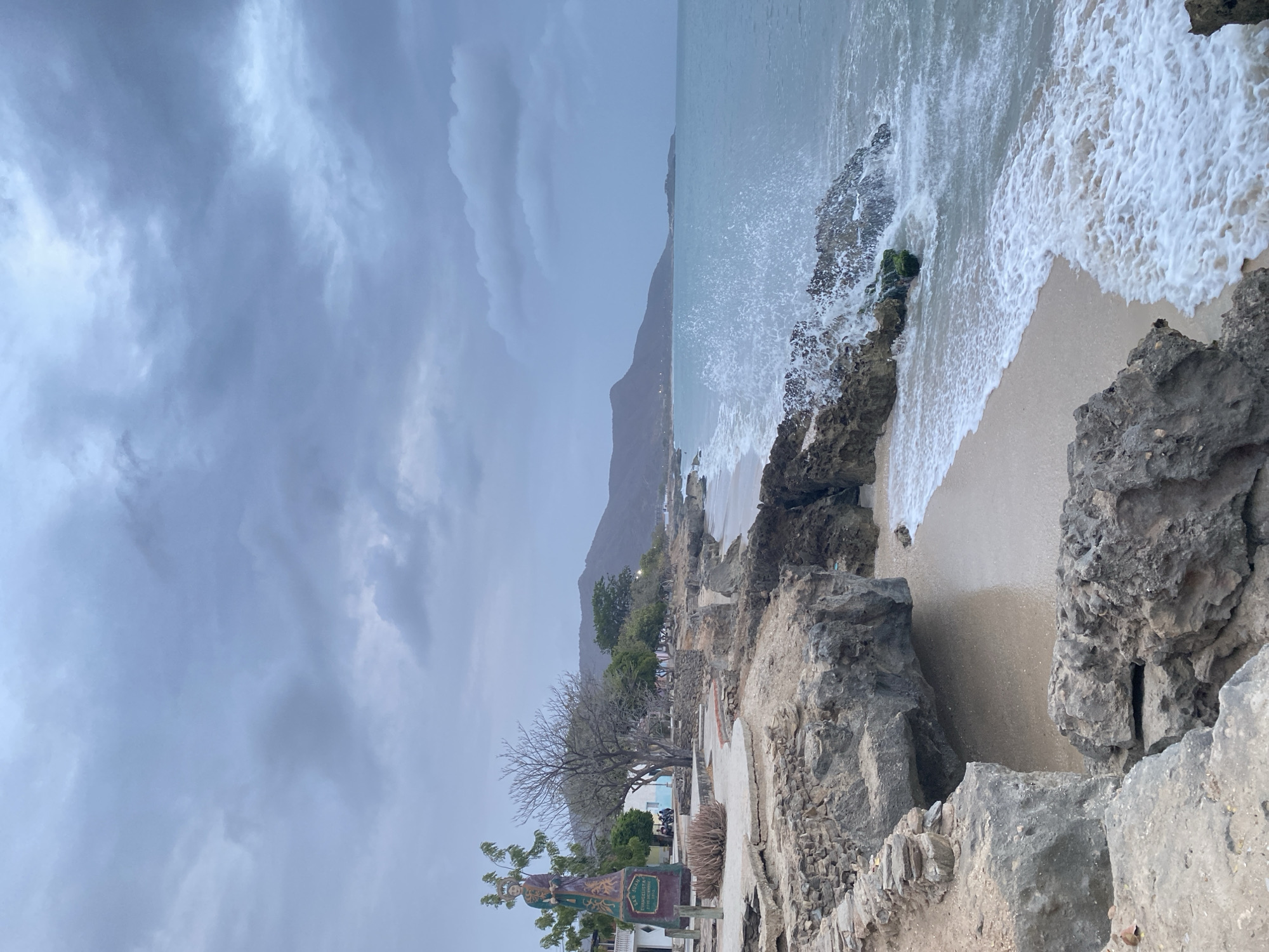 The image captures a coastal scene in Juan Griego, Venezuela, characterized by a rocky shoreline, a beach, and a body of water, likely the Caribbean Sea. A mountain looms in the distance, partially obscured by a cloudy, overcast sky.
In the foreground, there's a rocky outcrop, with visible signs of weathering and erosion. A sandy beach is visible between the rocks and the water's edge. Waves are crashing against the rocks, creating white foam and splashes.
In the mid-ground, there is some sparse vegetation along the coastal area, including trees and shrubs. There's a walkway or path that seems to be adjacent to a cemetery, indicated by gravestones and memorial structures. A religious statue is also noticeable.
The background is dominated by a mountain range that stretches along the coastline. The sky is heavily overcast with dark, layered clouds suggesting an impending storm or recent rainfall.
The scene is presented in a vertical orientation.