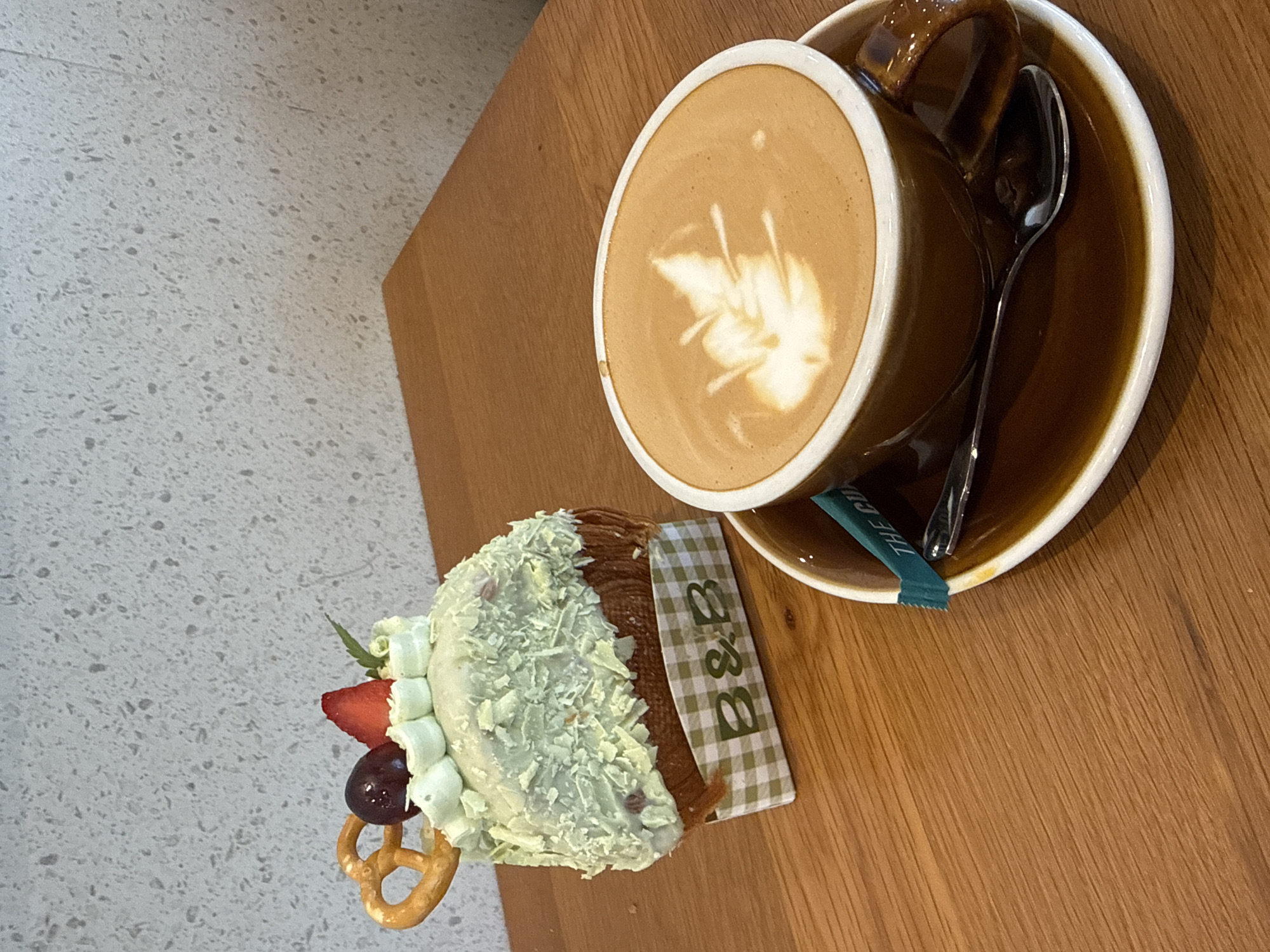 A top-down shot shows a table with a cappuccino in a brown mug on a saucer with a spoon and a sugar packet, and a piece of pastry on a checkered paper. The pastry is decorated with white frosting, a strawberry, a grape, and a pretzel. The background is a wood-grain table and a wall with a textured surface. The location is Unknown, Unknown.
