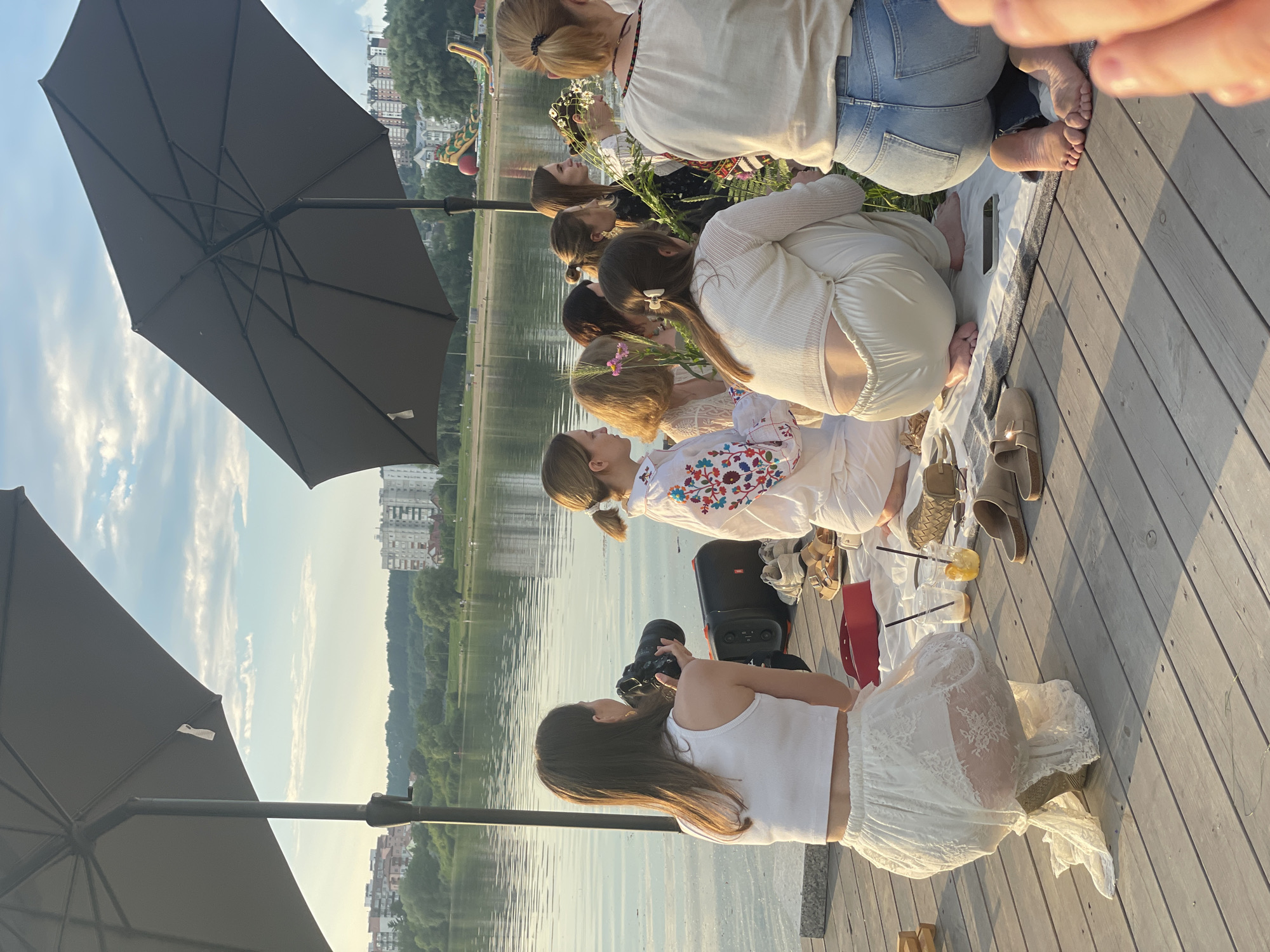 A group of women are having a picnic on a wooden pier next to a body of water, likely the Southern Bug River in Khmelnytskyi, Ukraine. The women are casually dressed, with some wearing embroidered blouses. One woman is holding a camera, seemingly taking photos of the group. There are picnic blankets, drinks, and shoes scattered on the pier. In the background, there are buildings and trees lining the opposite bank of the river, and a cloudy sky with the sun beginning to set.