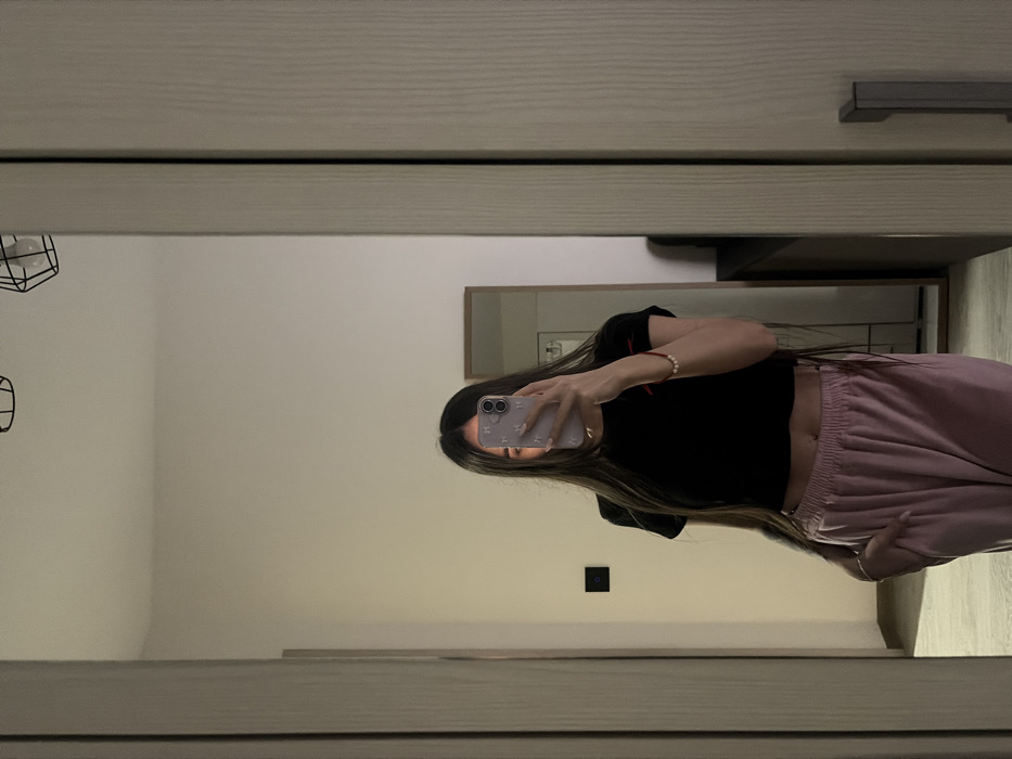 A female individual is depicted in an indoor setting, reflected in a large mirror. The exact location within Novosaratovka, Russia, cannot be determined from the image. The individual, whose face is obscured by a light purple smartphone with a patterned case, is wearing a black short-sleeved crop top revealing her abdomen and light pink sweatpants. Her long, dark hair extends over her shoulders and partially covers her chest. Her left hand is placed in her pants pocket, while her right hand holds the smartphone, positioned to capture her reflection. A red string bracelet with a white bead is visible on her right wrist. The reflected environment features light-colored walls and flooring, with two black geometric pendant light fixtures visible on the ceiling. A black rectangular light switch is affixed to the wall on the left side of the reflection. Further into the background, another rectangular mirror with a wooden frame is visible, reflecting a portion of the room, alongside a dark piece of furniture. The main mirror appears to be part of a door or closet, indicated by a handle on its right side and a vertical textured panel adjacent to it.