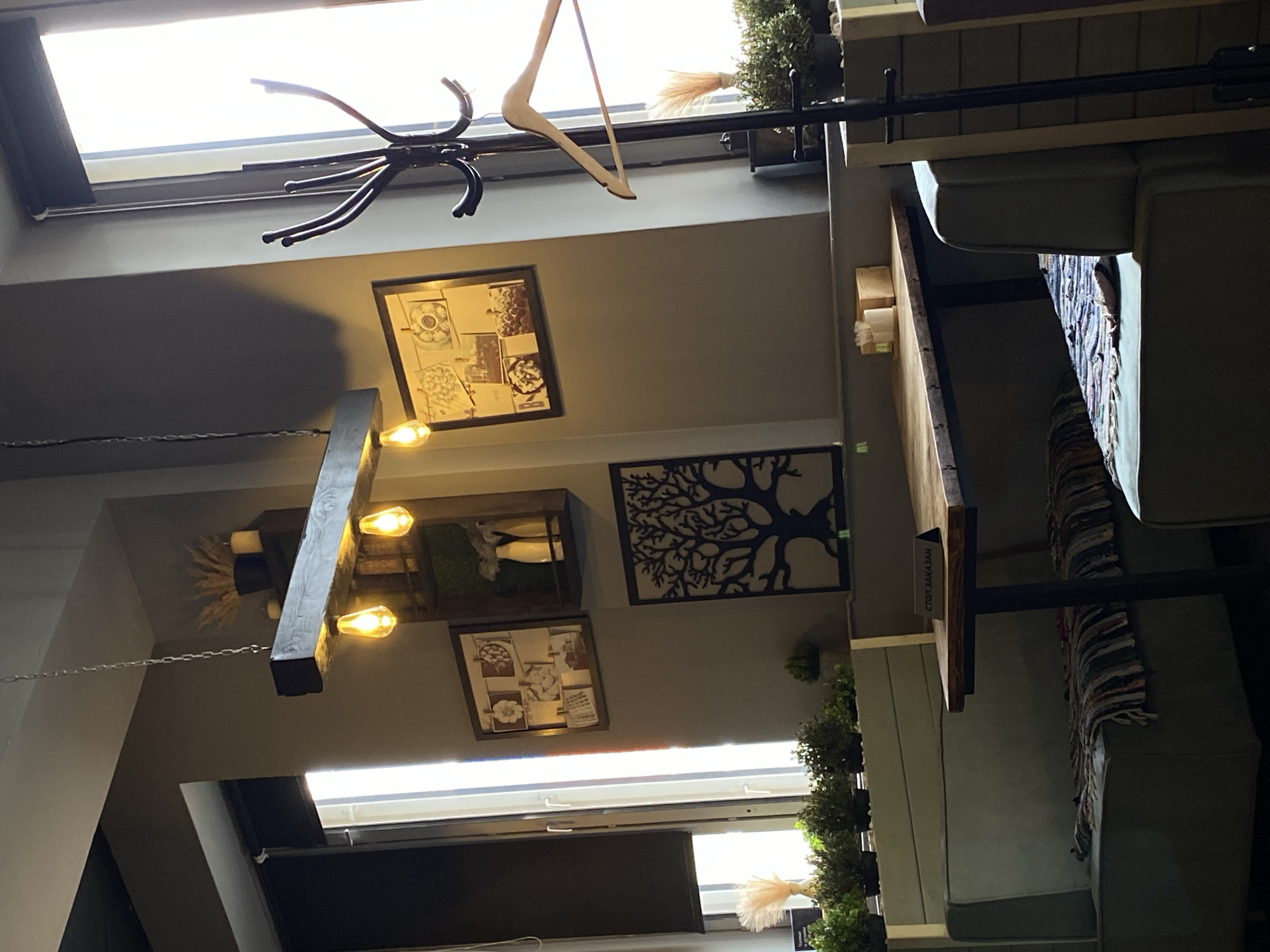The image depicts an interior shot of a room with a modern aesthetic. The ceiling features a combination of recessed lighting and natural light from skylights. Various decorative elements are present, including framed artwork, a tree silhouette design, and a lighting fixture with exposed bulbs. A clothes hanger and a coat rack are also visible near the top of the frame. On the right, a banquette with cushions is situated against a paneled wall adorned with greenery.
The location appears to be a restaurant or cafe in Votkinsk, Russia, judging by the sign "СТОЛ ЗАКАЗАН" (Table Reserved in Russian) on the banquette table.