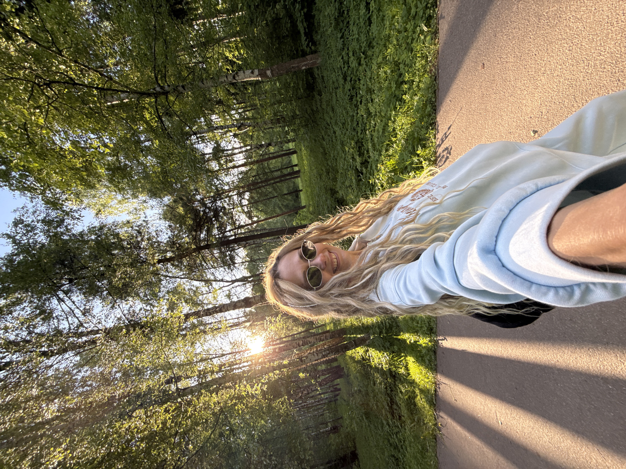 A blonde woman wearing sunglasses and a blue sweatshirt is taking a selfie. She is on a path in a park, with trees to her left and a strip of grass between the path and the trees. Sunlight is visible through the trees. The location is Шебунино, Russia.