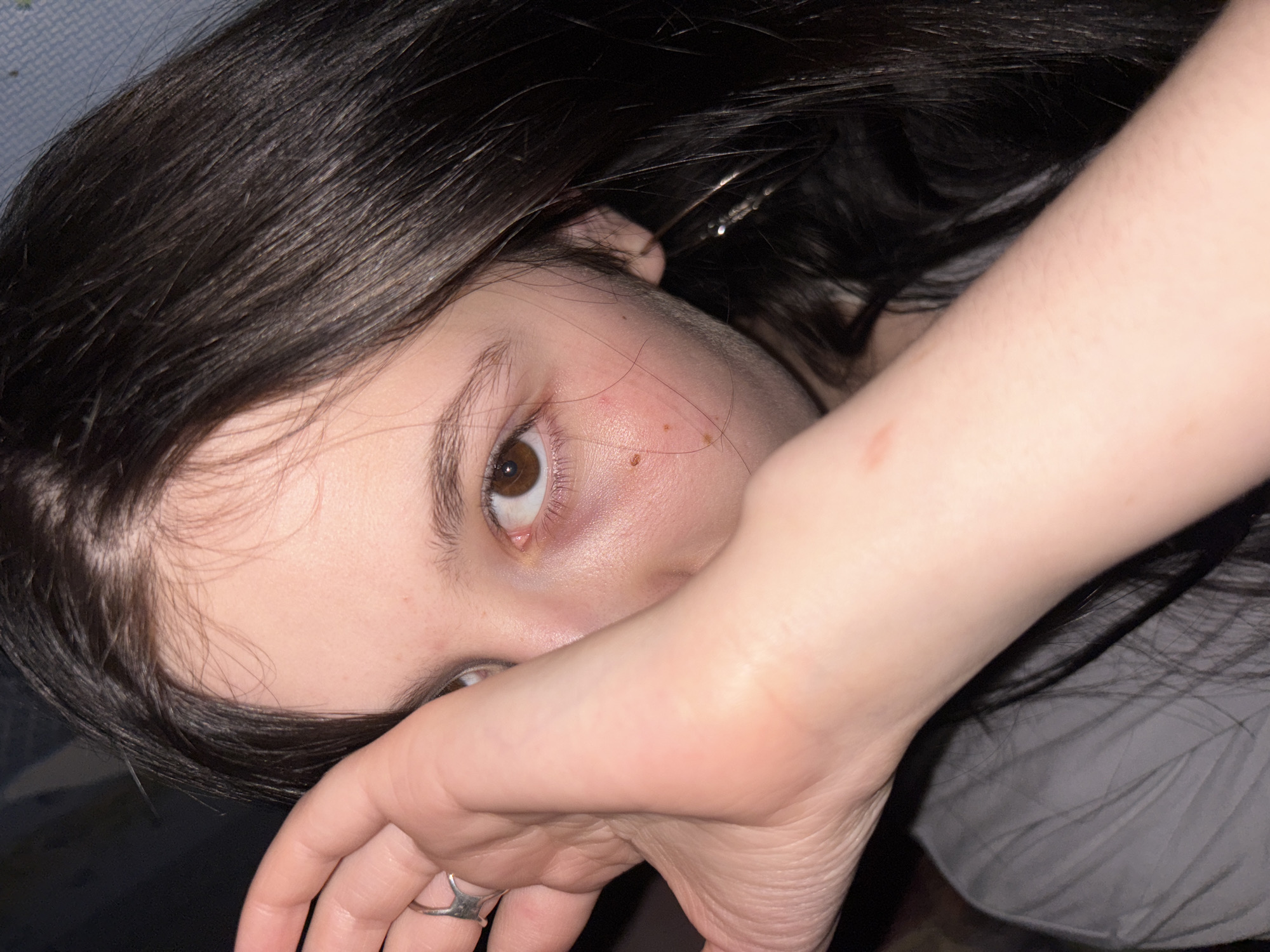 A close-up, angled shot of a fair-skinned person with dark brown hair. The person's face is partially obscured by their hand and arm. Only one eye is fully visible, it is brown. A silver ring is on one of the fingers covering the face. The background is out of focus, with a mottled blue-gray texture visible in the upper left corner. The person appears to be indoors. There is no indication of Фоки, Russia or a city environment.