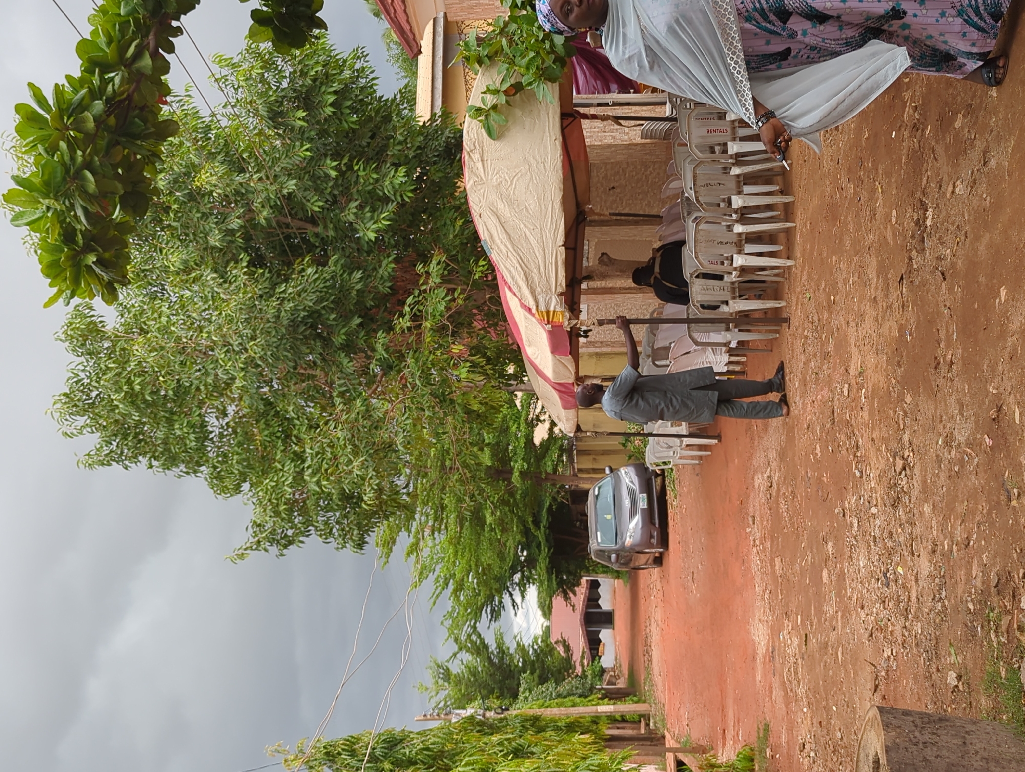 The image depicts an outdoor scene in Unguwar Rimi, Nigeria, characterized by reddish-brown dirt ground and an overcast, grey sky. Lush green trees, with varied foliage, dominate the left side of the frame, with power lines visible extending across the top and mid-ground. In the background, buildings with light-colored walls and reddish-brown roofs are partially visible.
On the right side, a temporary canopy or awning, beige with red stripes, is set up, supported by poles. Beneath it, numerous white plastic chairs are arranged and stacked, indicating preparations for an event or gathering. The word "RENTALS" is clearly visible printed on the backs of several chairs, suggesting they were hired for the occasion.
Several individuals are present. On the far right, a woman wearing a light-colored head covering (hijab) and a patterned dress stands, looking towards the camera. She holds a dark object, possibly a pen, in her hand. Closer to the center, under the canopy, a man in a dark grey suit or uniform is standing, holding an upright pole, possibly involved in the setup or organization of the event. Other figures are partially discernible under the canopy and among the chairs in the background.
A dark-colored minivan or car is parked on the dirt path in the mid-ground. A large, cut log or tree trunk is partially visible in the bottom right corner. The overall scene suggests a community gathering or outdoor event taking place during the daytime, under cloudy weather conditions.
