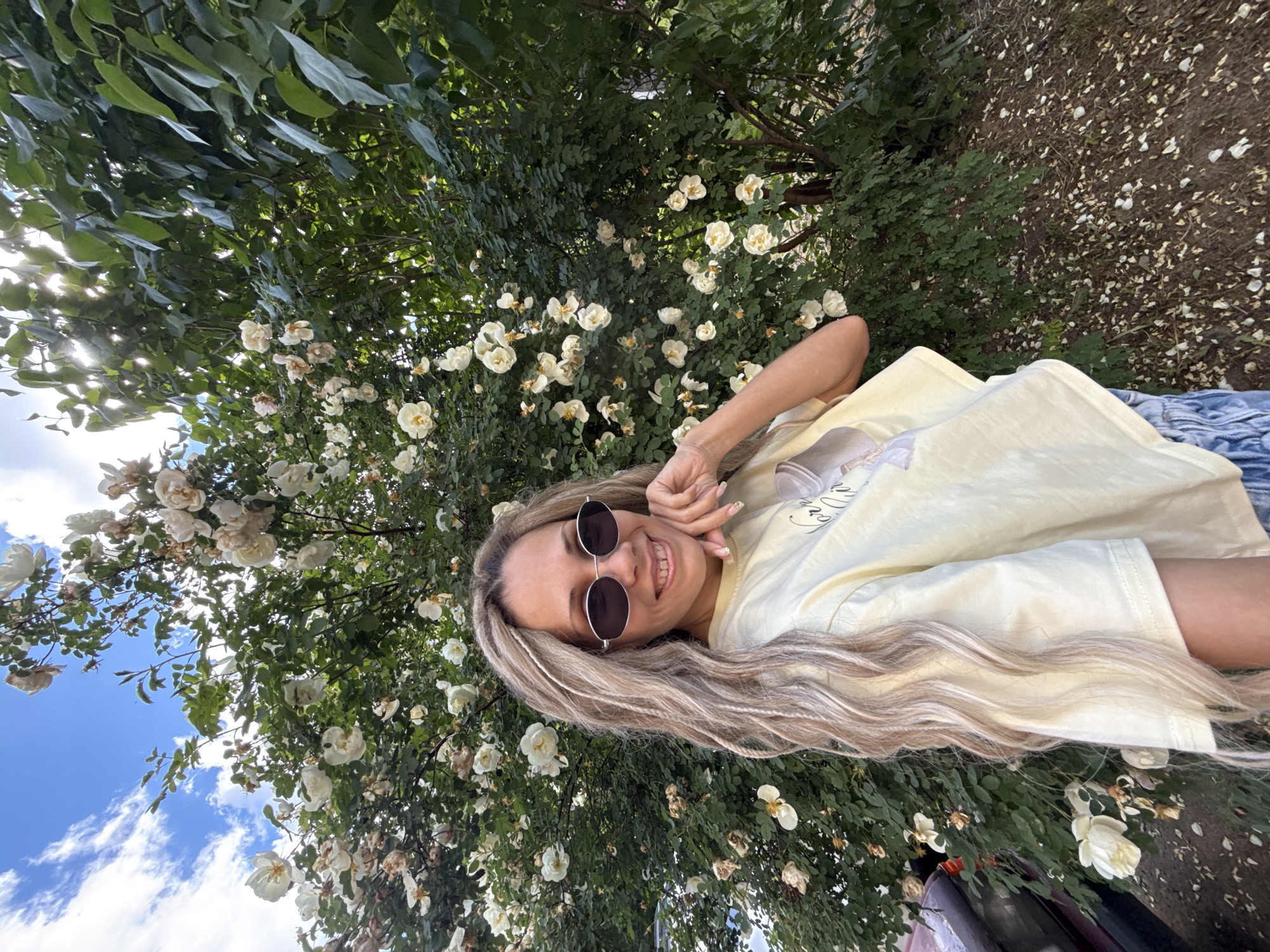 The image, oriented sideways, displays a cheerful young woman lying amidst a profusion of white roses and green foliage. Her head is positioned towards the left of the frame, and she is smiling brightly, looking directly at the camera. She has long, wavy blonde hair and wears round, dark sunglasses. A light yellow t-shirt, possibly featuring the text "Sunny Way," is visible on her torso. Her right hand is gently resting near her face.
The background is dominated by dense green leaves and numerous white roses, some fully bloomed and others with fading petals. Bright sunlight filters through the foliage from the upper left, creating highlights. To the left, a clear blue sky with fluffy white clouds suggests a bright day. The right side of the image shows the ground, covered with scattered dry leaves and fallen white petals, indicating a natural, outdoor setting. The scene evokes a feeling of relaxed enjoyment within a beautiful garden or park in Rusanovo, Russia.