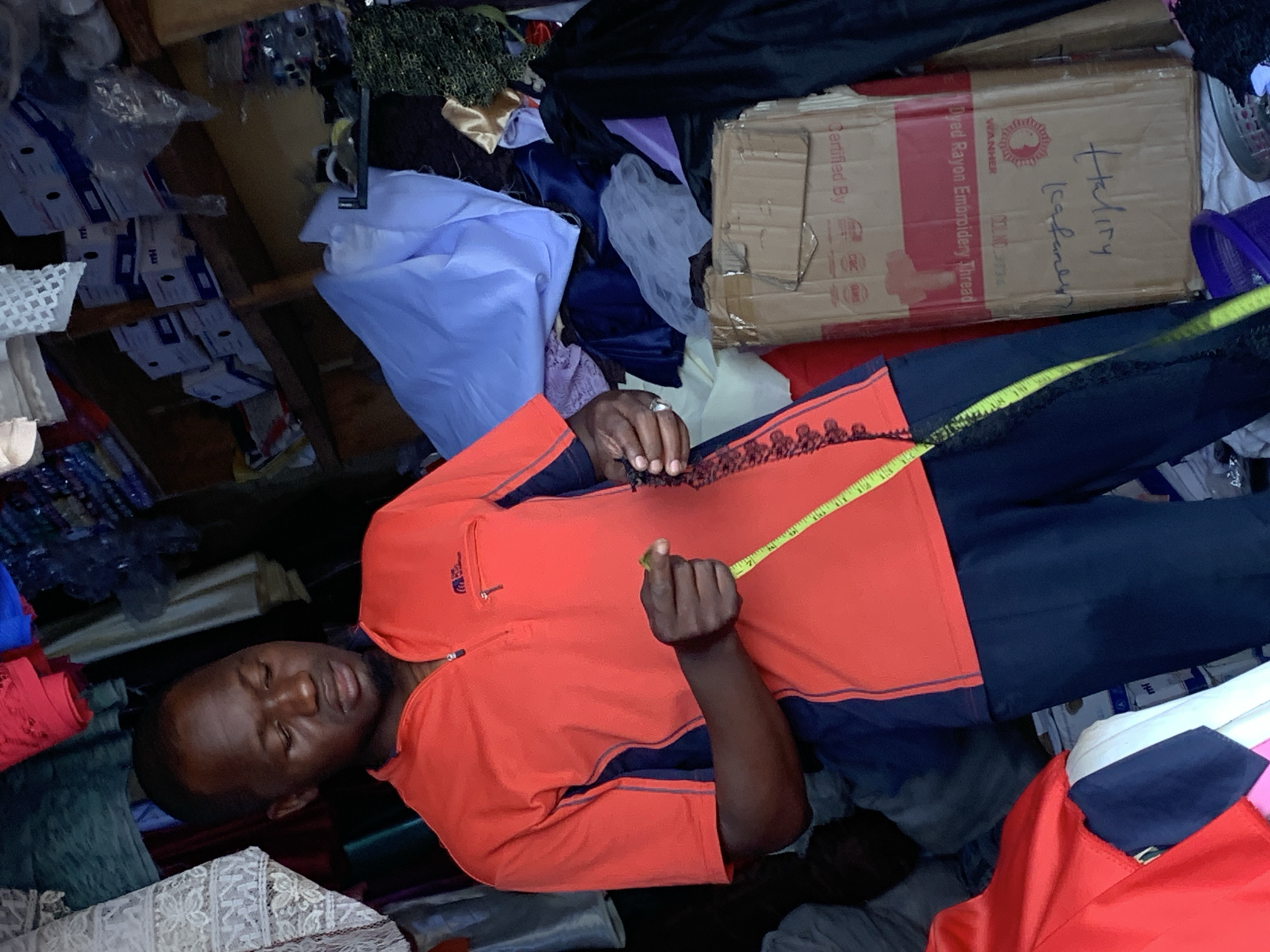 The image shows a man in an orange and navy blue shirt holding a yellow measuring tape. He is surrounded by various textiles and fabrics, including blue, white, black, and other colored materials. A cardboard box labeled "Dyed Rayon Embroidery Thread" and "Certified By" is visible. The environment suggests a workshop or small business, given the presence of fabrics and sewing-related items. This appears to be a small business setting in Kafanchan B, Nigeria.