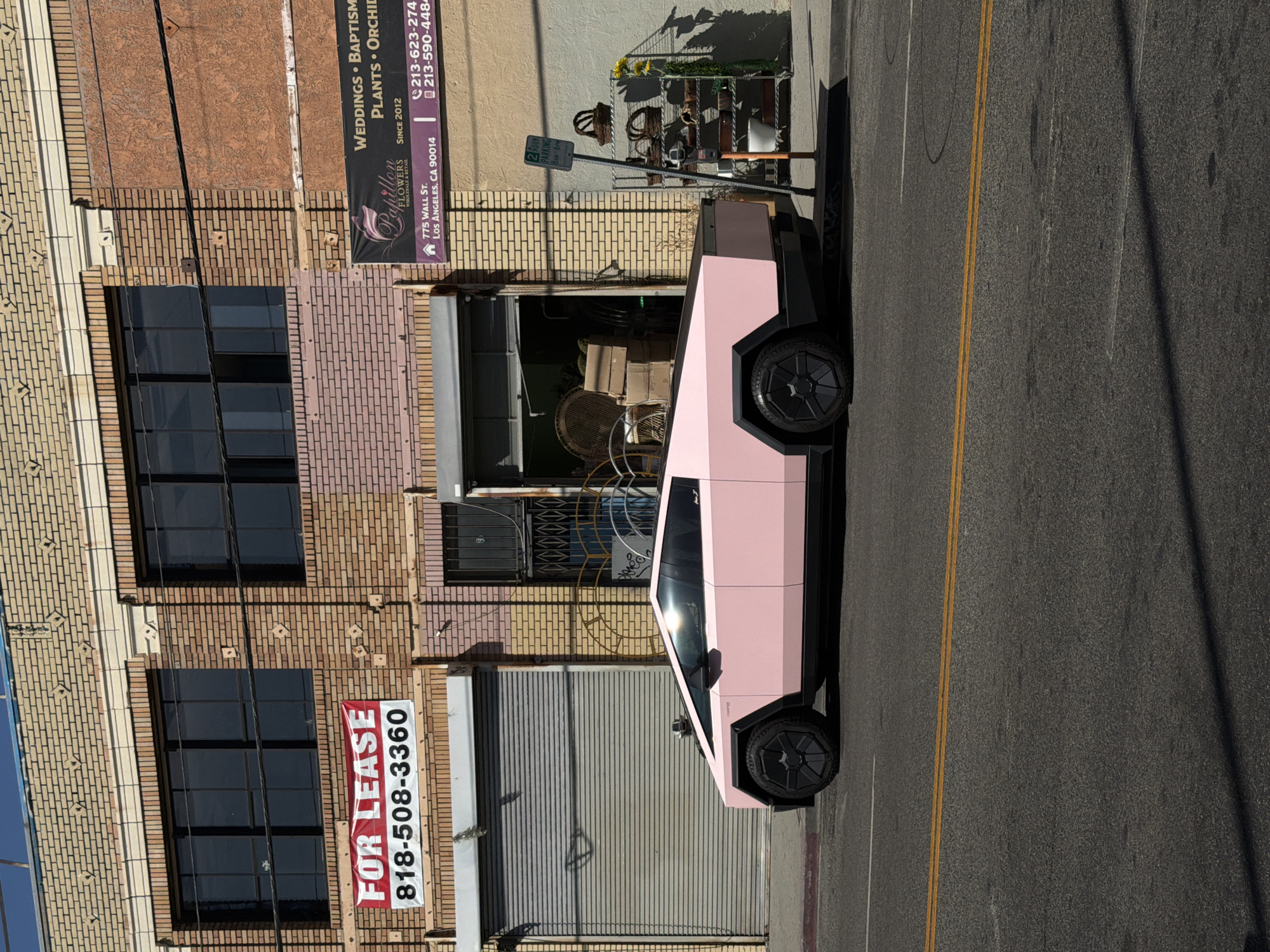 A pink Tesla Cybertruck is parked on the street in Los Angeles, California, United States of America. The Cybertruck is parked adjacent to a brick building with a "FOR LEASE" sign prominently displayed. A flower shop named "Papillon Flowers" is visible next to the building, indicating the location as 775 Wall Street, Los Angeles, CA 90014. A 2-hour parking sign is also present. The street has double yellow lines. There is a store front filled with props behind the cybertruck.