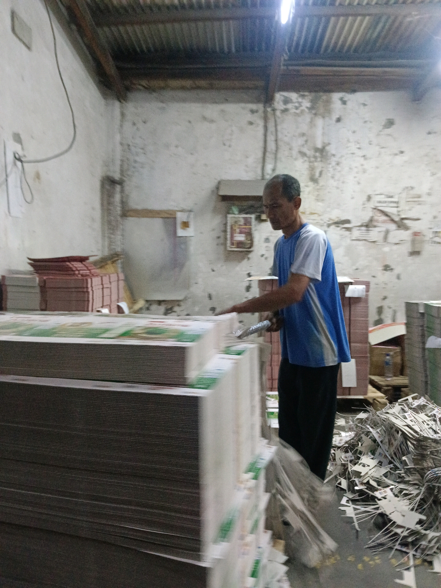 In Bringinbendo, Indonesia, a man is seen in what appears to be a printing or paper processing facility. The environment is utilitarian, with a corrugated metal roof and stark, textured walls that show signs of age and discoloration. A single light fixture illuminates the space from above.
The man, wearing a blue and white t-shirt and dark pants, is actively engaged in his work. He is handling stacks of paper or printed materials, some of which are precariously balanced. His right hand is holding what appears to be a roll of tape or some other binding material.
In the foreground, large stacks of paper are prominently displayed, creating a sense of scale and the volume of material being processed. To the right, a pile of discarded paper scraps or trim is visible, indicating the byproduct of the operation. In the background, more stacks of what look like boxed paper products or bound volumes are organized. The overall impression is of a busy, working environment dedicated to the handling and processing of paper goods.