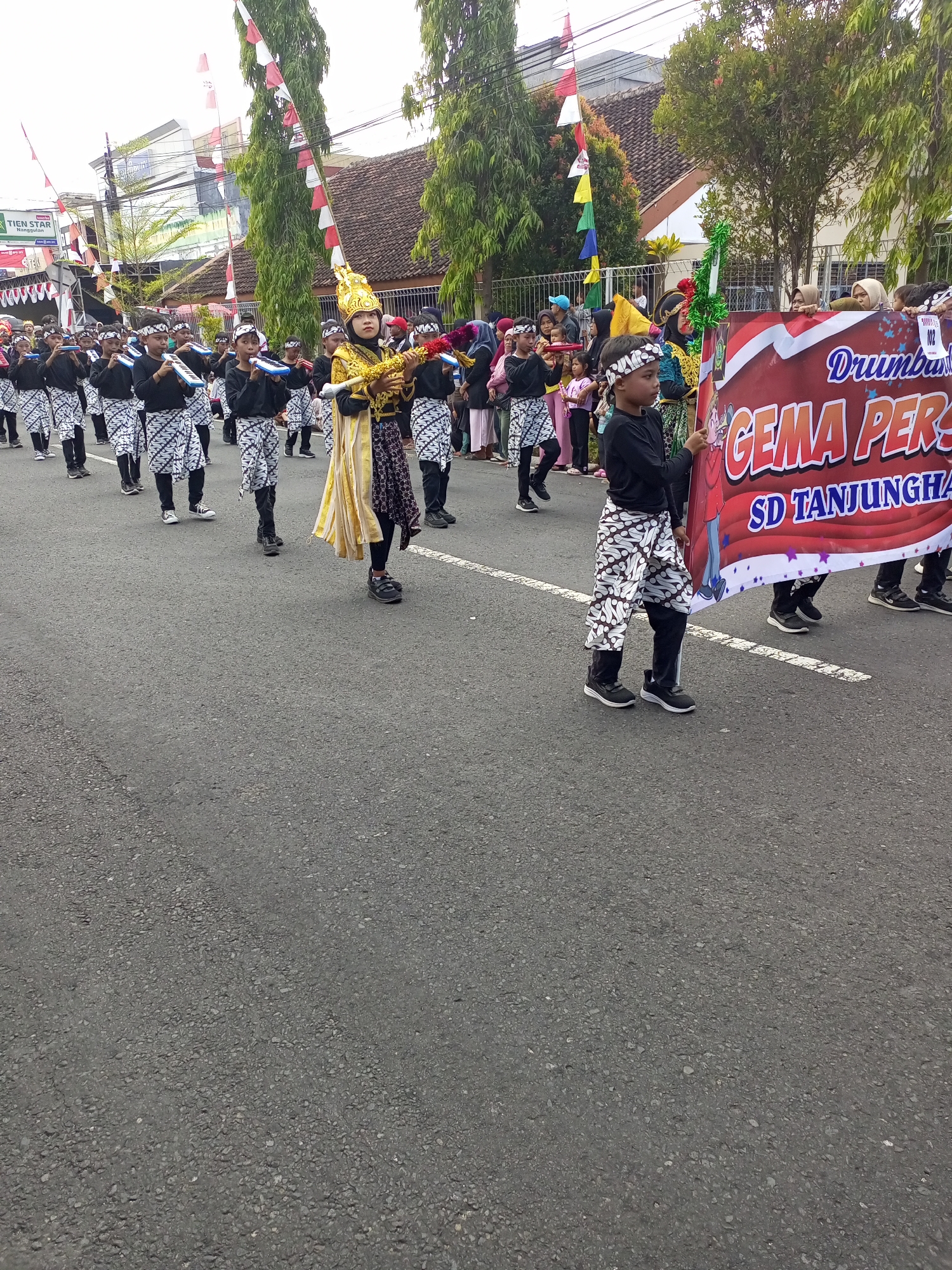 In Nanggulan, Indonesia, a parade is underway on a paved road. The central focus is a group of children dressed in traditional attire, participating in a musical performance. One child in the foreground, adorned with a golden headdress and ornate yellow and gold clothing, holds a brass instrument. Behind them, a line of children, also in traditional black tops and patterned pants, play similar instruments. To the right, another child holds a large red banner with white lettering that reads "GEMA PERS... SD TANJUNGHA." The parade is lined by spectators and decorated with red and white flags and green streamers. The environment suggests a town street with buildings and trees visible in the background.
