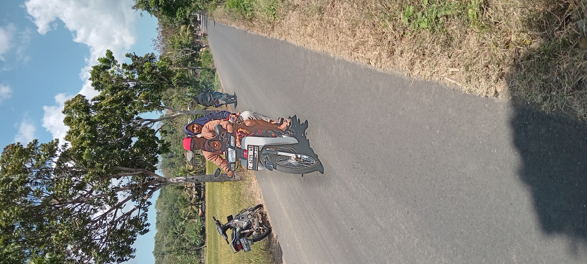 In Paledah, Indonesia, on a paved road bordered by dry grass and a rice field, a family of three rides a motorcycle. The driver, a man wearing a red cap and brown shirt, is in the front, followed by a woman in a blue hijab and a child wearing a pink headscarf in the middle. The motorcycle is white and has a license plate reading "Z 6215 HA." In the background, another motorcycle with a person wearing black is visible further down the road. Trees with green foliage line the side of the road, and a cluster of houses can be seen amongst them. The sky is blue with scattered white clouds. A second motorcycle is parked on the left side of the road.